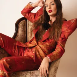 Portret kobiety w czerwonym wzorzystym garniturze, siedzącej w fotelu w stylu vintage, z mocnym makijażem ust i rozpuszczonymi włosami, pozującej w studiu.