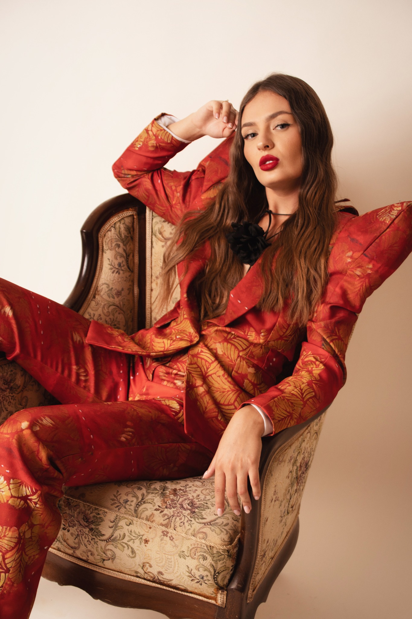Portret kobiety w czerwonym wzorzystym garniturze, siedzącej w fotelu w stylu vintage, z mocnym makijażem ust i rozpuszczonymi włosami, pozującej w studiu.