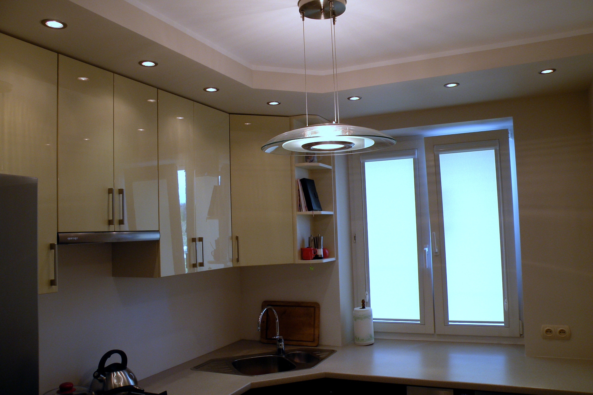 Jasna kuchnia z połyskującymi frontami szafek, nowoczesnym oświetleniem sufitowym i okapem nad płytą grzewczą. Widoczny zlew narożny, czajnik i deska do krojenia.