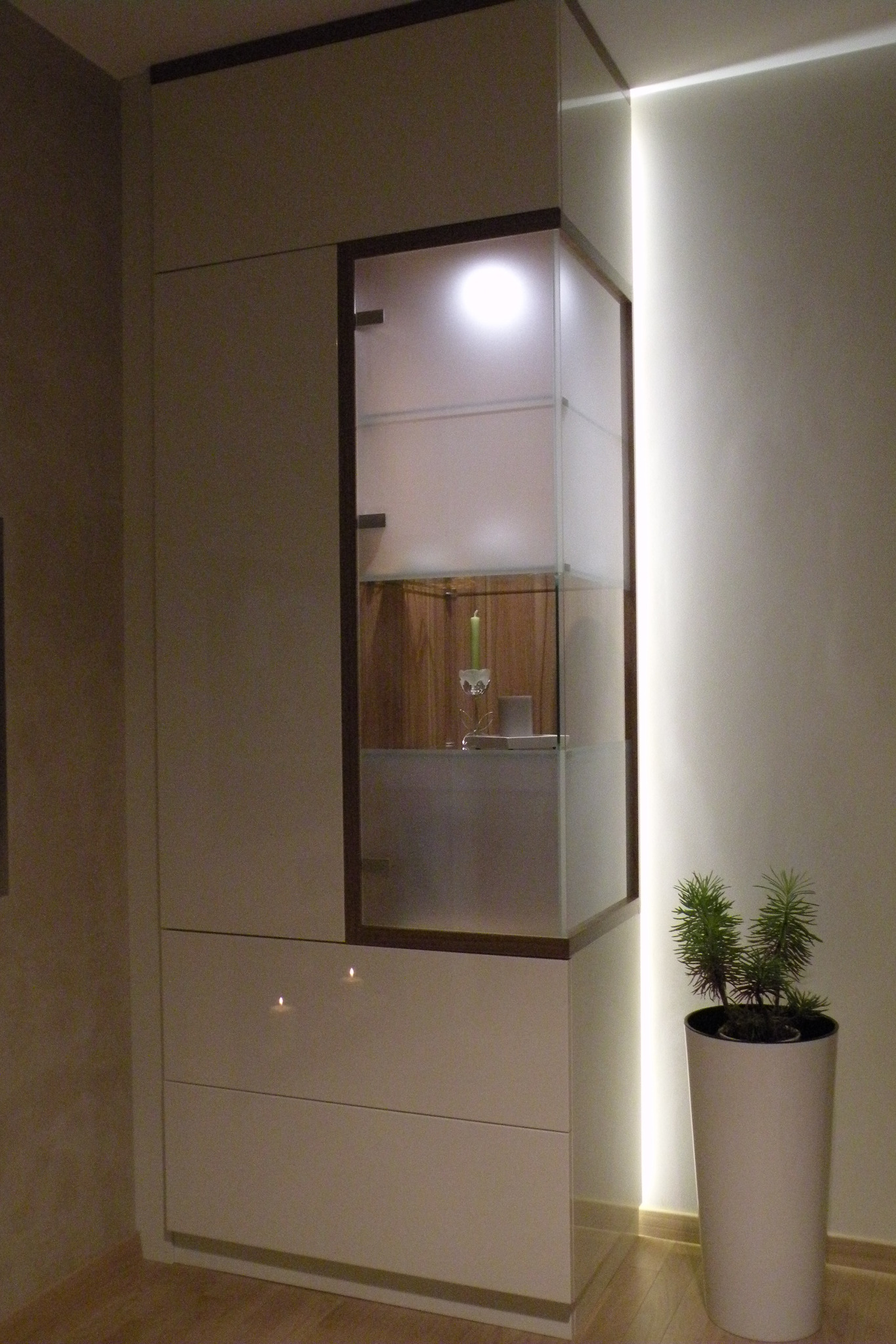 Biała szafa na wymiar z szufladami i przeszkloną witryną z podświetleniem LED, w której stoi świeca i dekoracje, obok doniczka z małą rośliną.