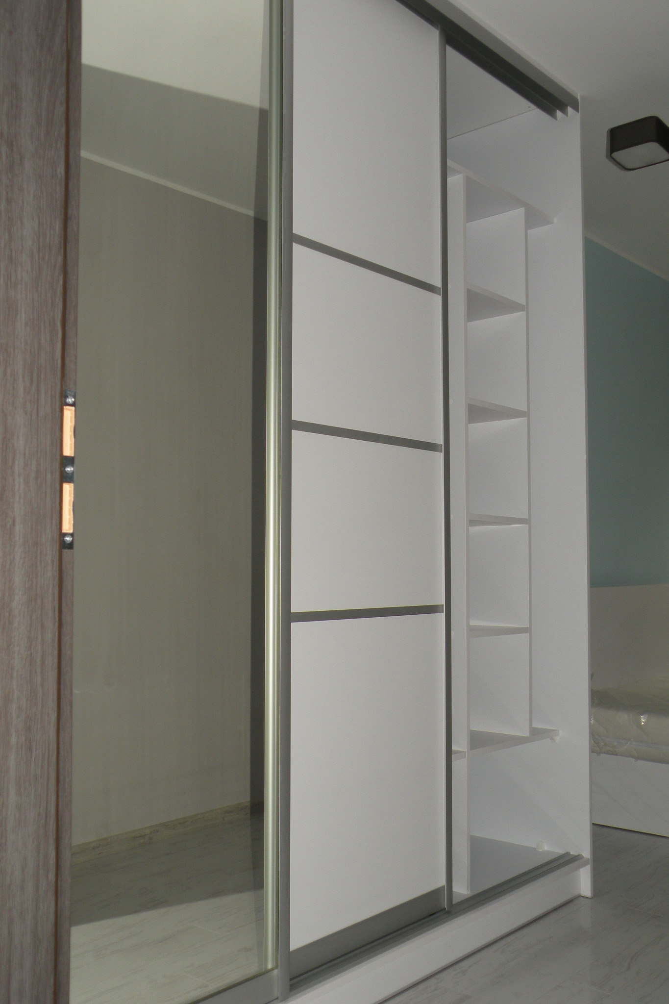 Biała szafa wnękowa z przesuwnymi drzwiami z lustrem, widoczne półki wewnętrzne, szare akcenty na drzwiach, zamontowana w pomieszczeniu z drewnianymi drzwiami.