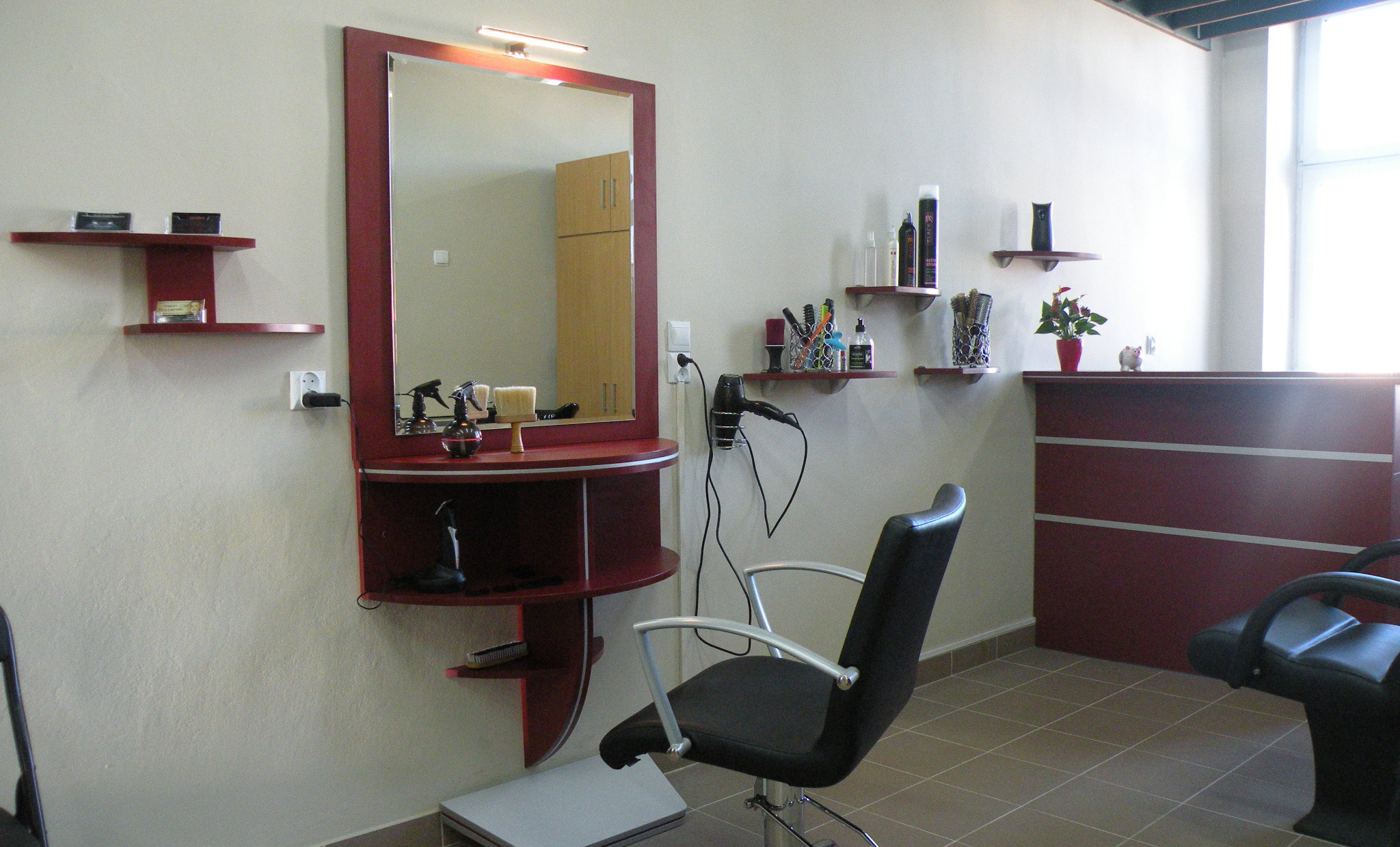 Stanowisko fryzjerskie z dużym lustrem w bordowej ramie, półkami na akcesoria i czarnym fotelem na srebrnej nodze, widok na wnętrze salonu z ladą recepcyjną.