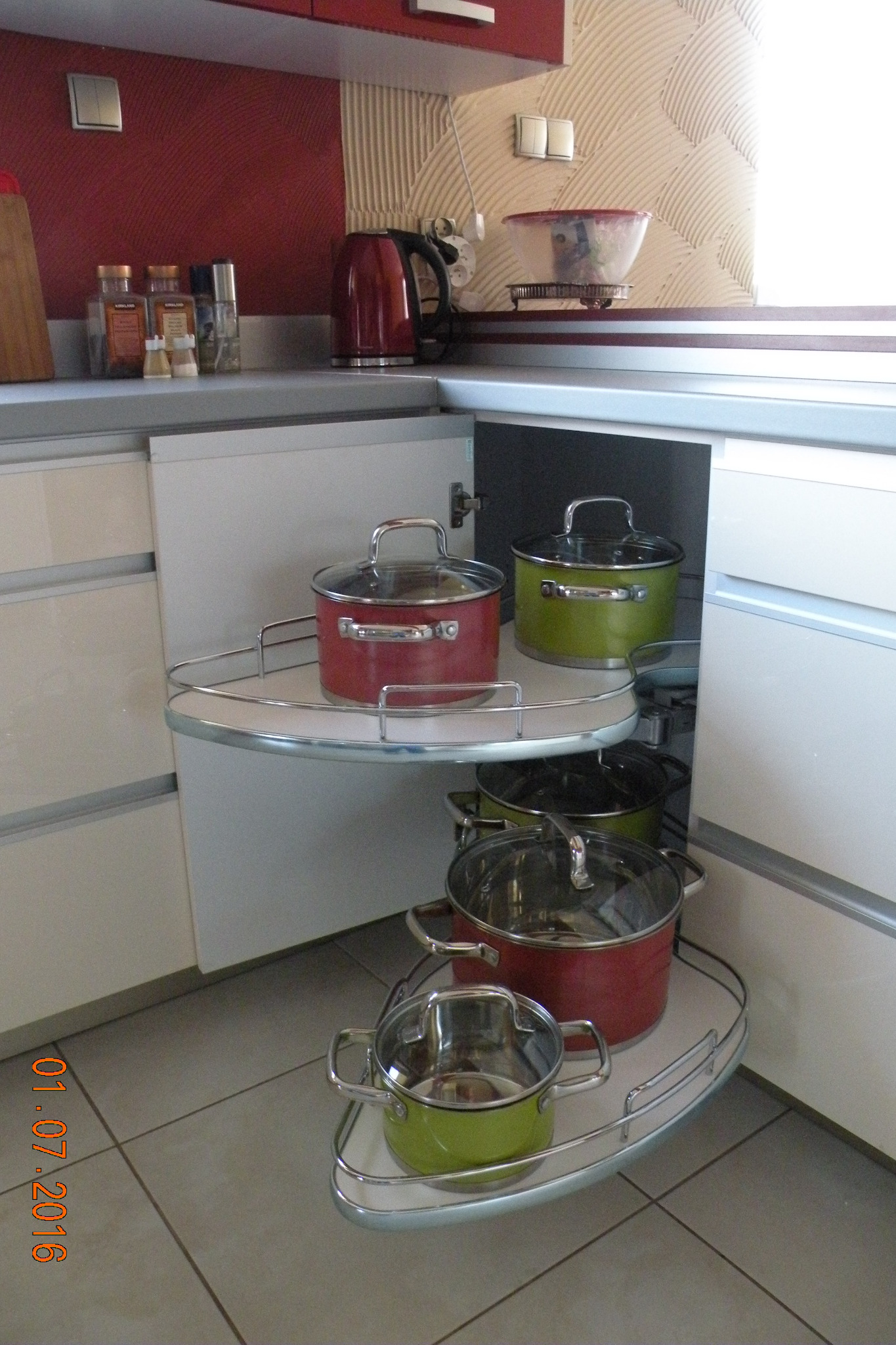Wewnętrzny narożnik szafki kuchennej z obrotowymi półkami, na których stoją garnki w kolorach czerwonym i zielonym.