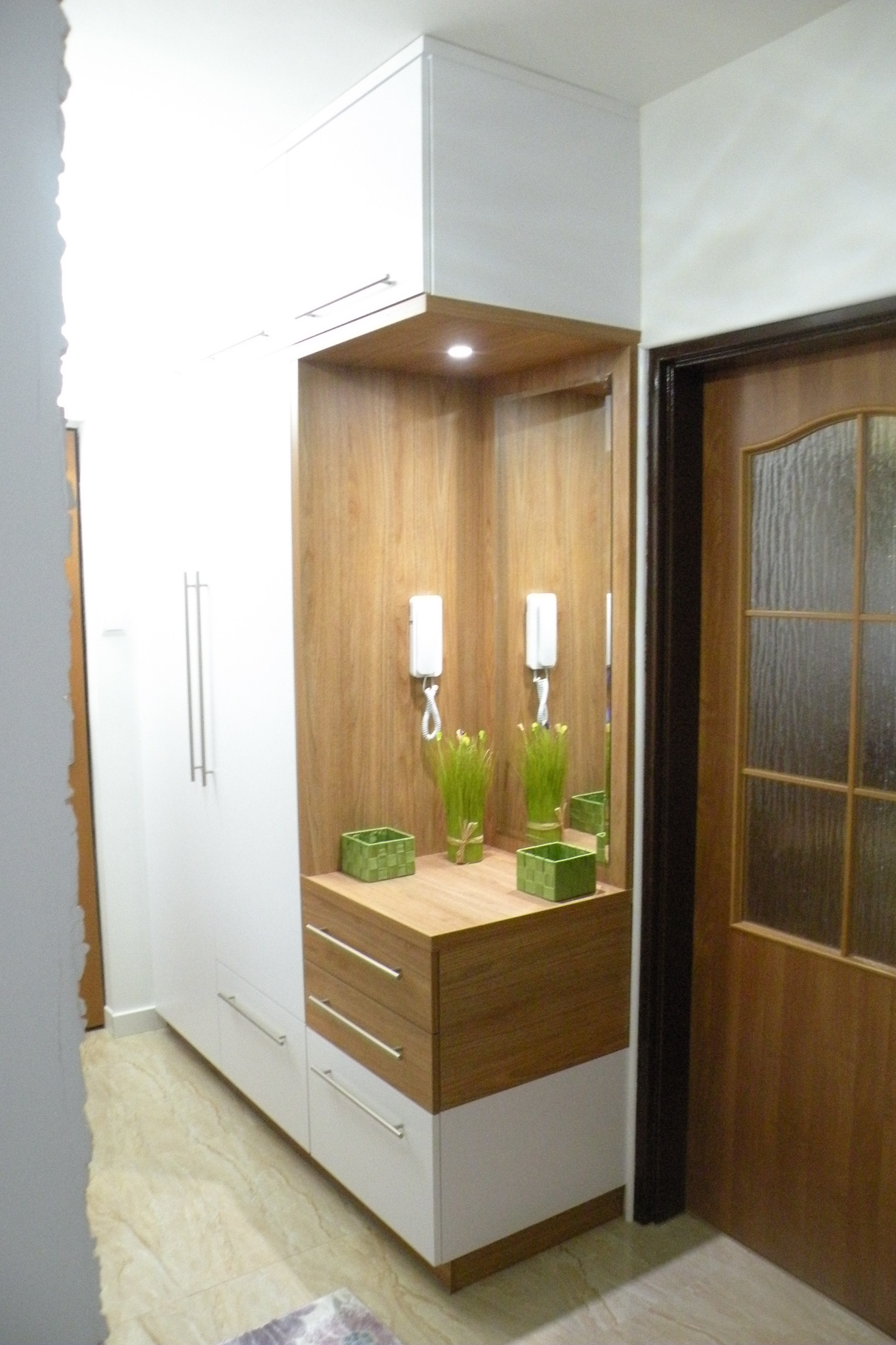 Biało-drewniana szafa wnękowa z szufladami i otwartą półką z podświetleniem, dekorowana zielonymi akcentami, w przedpokoju obok drzwi.