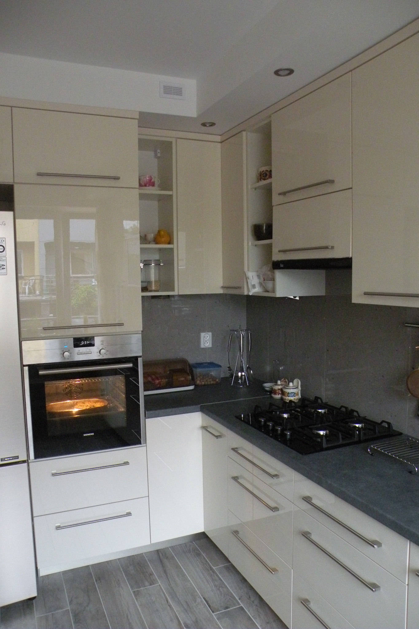 Jasna kuchnia z frontami na wysoki połysk, piekarnik zabudowany z piekącą się pizzą, szara ściana nad blatem i płyta gazowa.