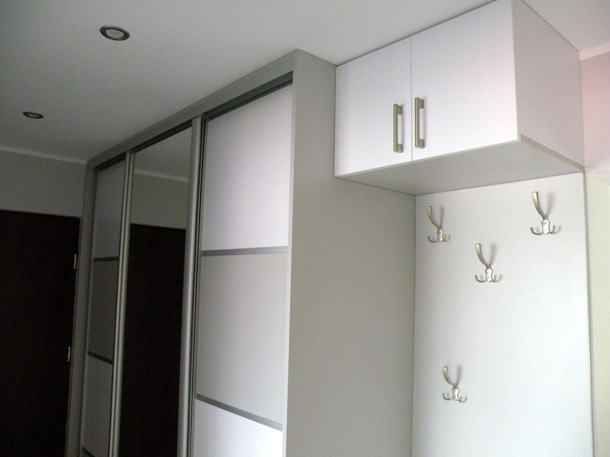 Biała szafa wnękowa z przesuwnymi drzwiami i szafką wiszącą z metalowymi uchwytami oraz wieszakami na ścianie, widziana z dołu.