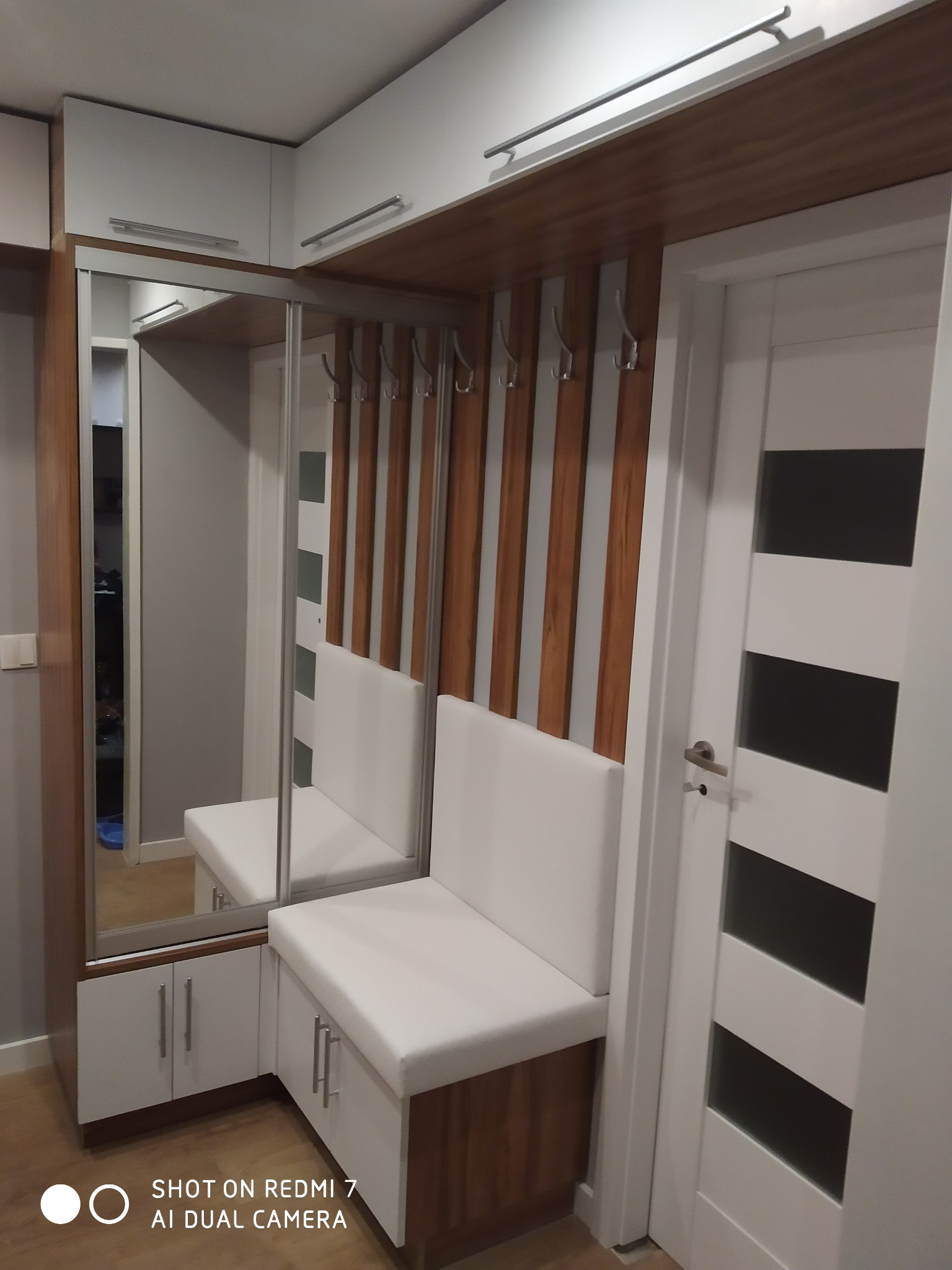 Przedpokój z siedziskiem i szafkami w zabudowie, lustro w aluminiowej ramie, wieszaki na ścianie z drewnianymi panelami, białe fronty szafek i siedziska, brązowe elementy wykończenia.