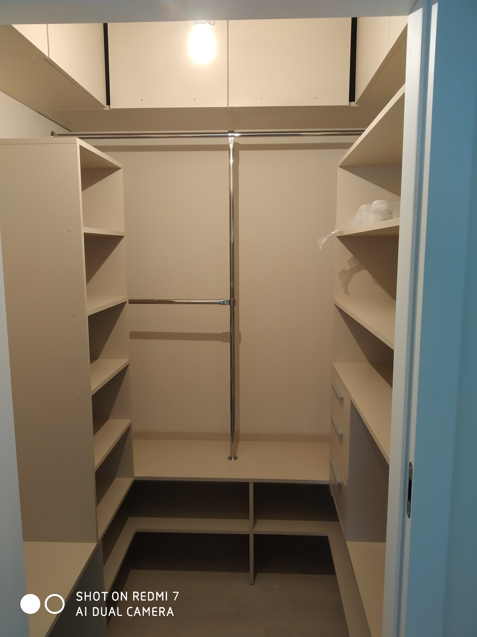 Wnętrze jasnej szafy wnękowej z półkami i drążkami na ubrania, widok z perspektywy wejścia, z plastikową folią ochronną na jednej z półek.