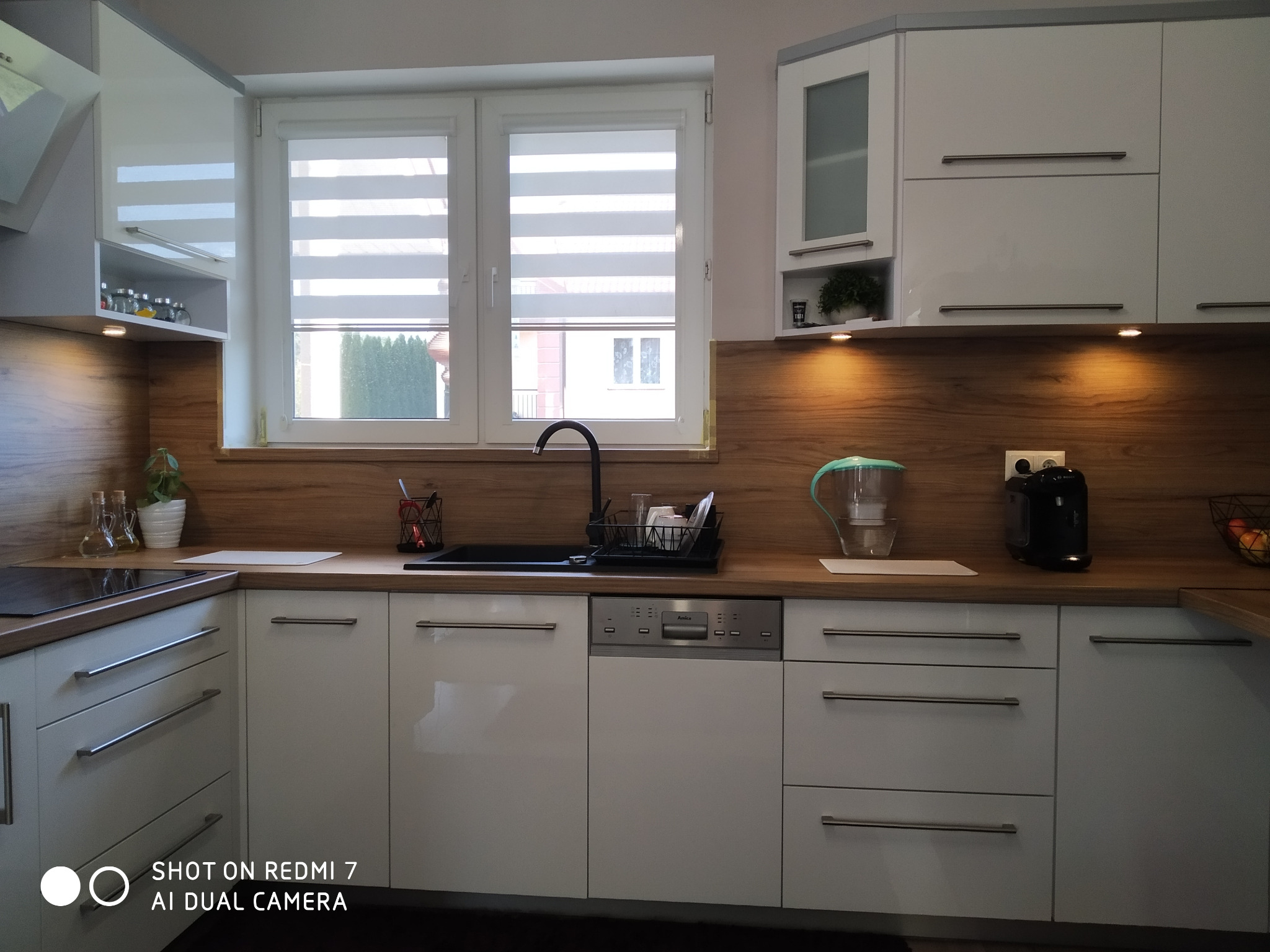 Jasna kuchnia z białymi, lakierowanymi szafkami, drewnianym blatem i roletami w oknie, widoczny czarny zlew i akcesoria kuchenne.