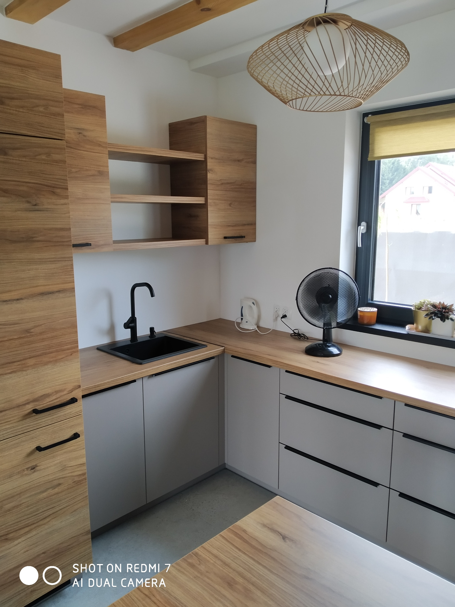 Nowoczesna kuchnia z drewnianymi szafkami górnymi i szarymi dolnymi, czarny zlew i bateria, drewniany blat, okno z roletą, wentylator na blacie, wisząca lampa z ażurowym kloszem.