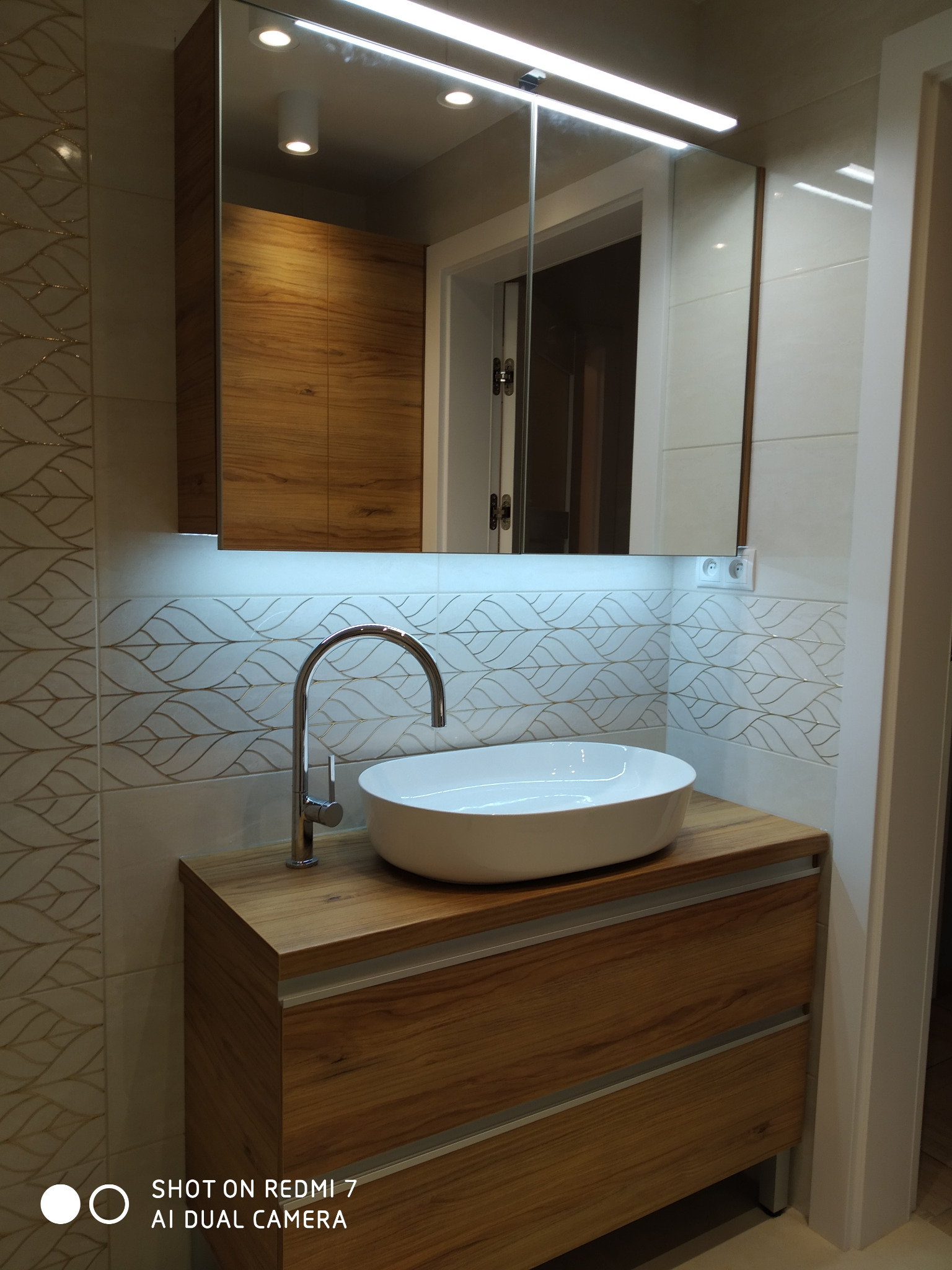 Nowoczesna łazienka z drewnianą szafką pod umywalką nablatową, lustrem z podświetleniem LED i dekoracyjnymi płytkami na ścianie.