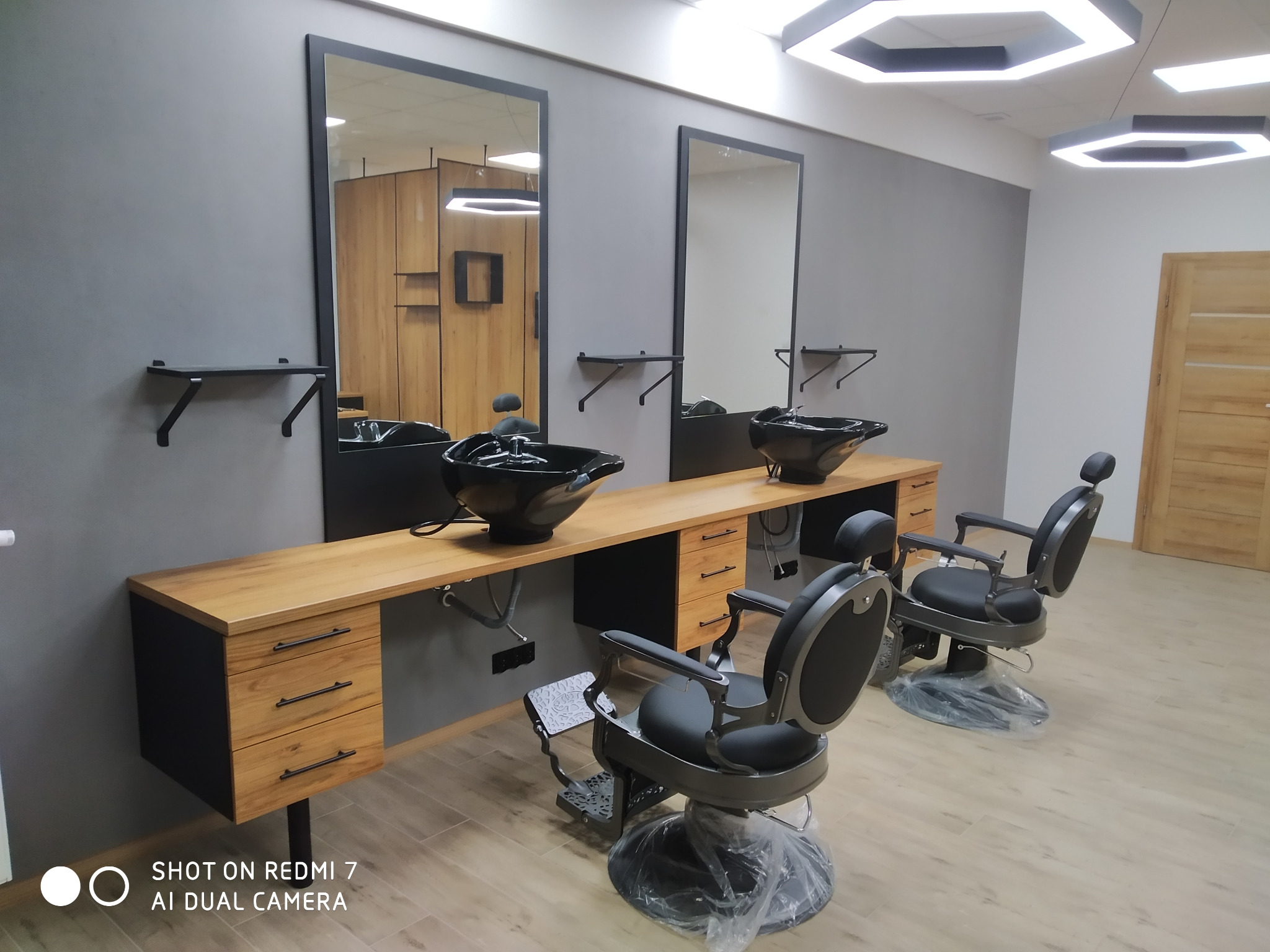Nowoczesne stanowiska fryzjerskie z drewnianymi blatami, czarnymi umywalkami i lustrami, oświetlone sześciokątnymi lampami.