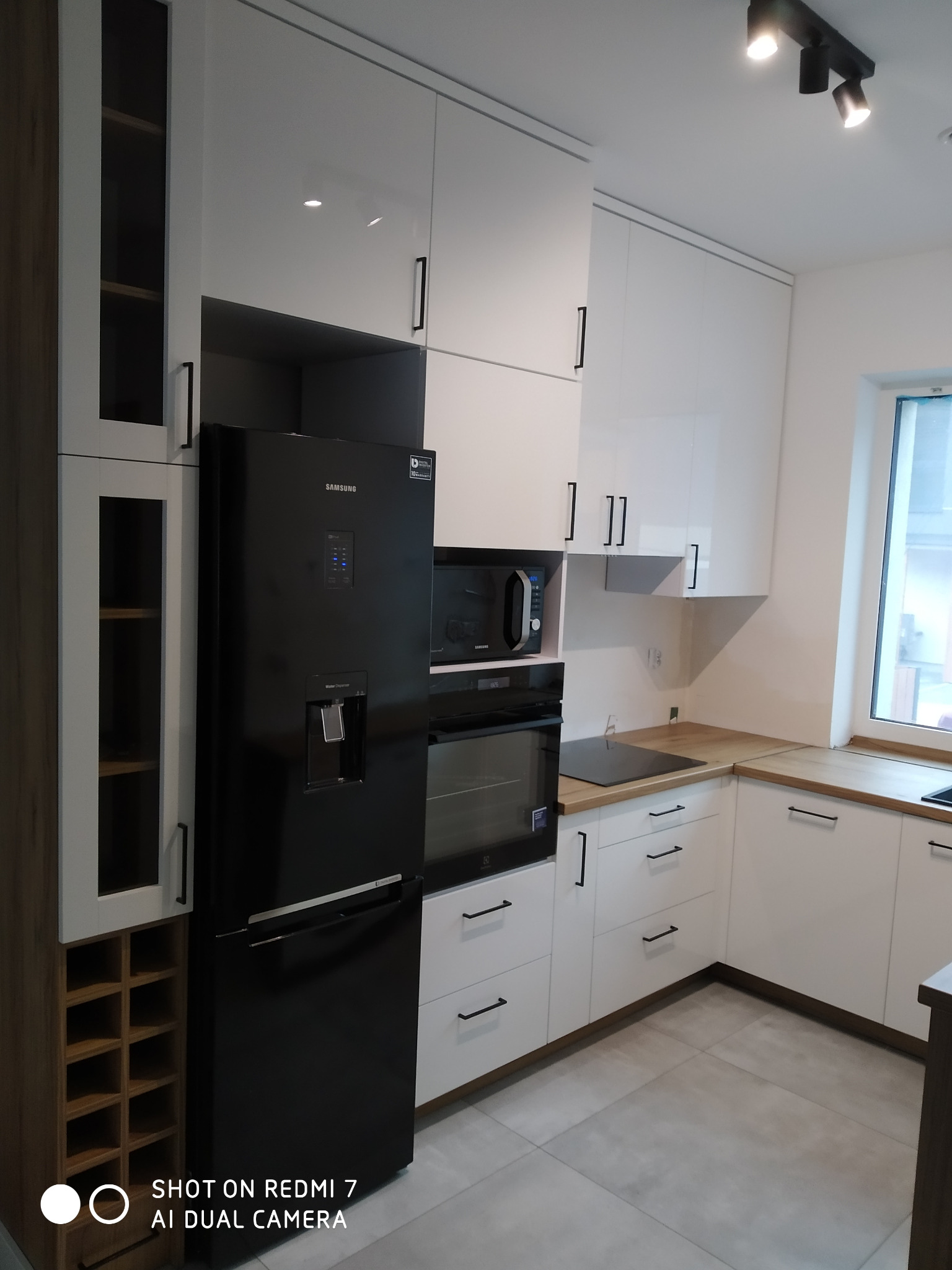 Nowoczesna kuchnia z białymi, błyszczącymi frontami szafek, czarnym sprzętem AGD (lodówka Samsung, piekarnik Electrolux, mikrofalówka) i drewnianym blatem. Szafka z półkami na wino po lewej stronie.