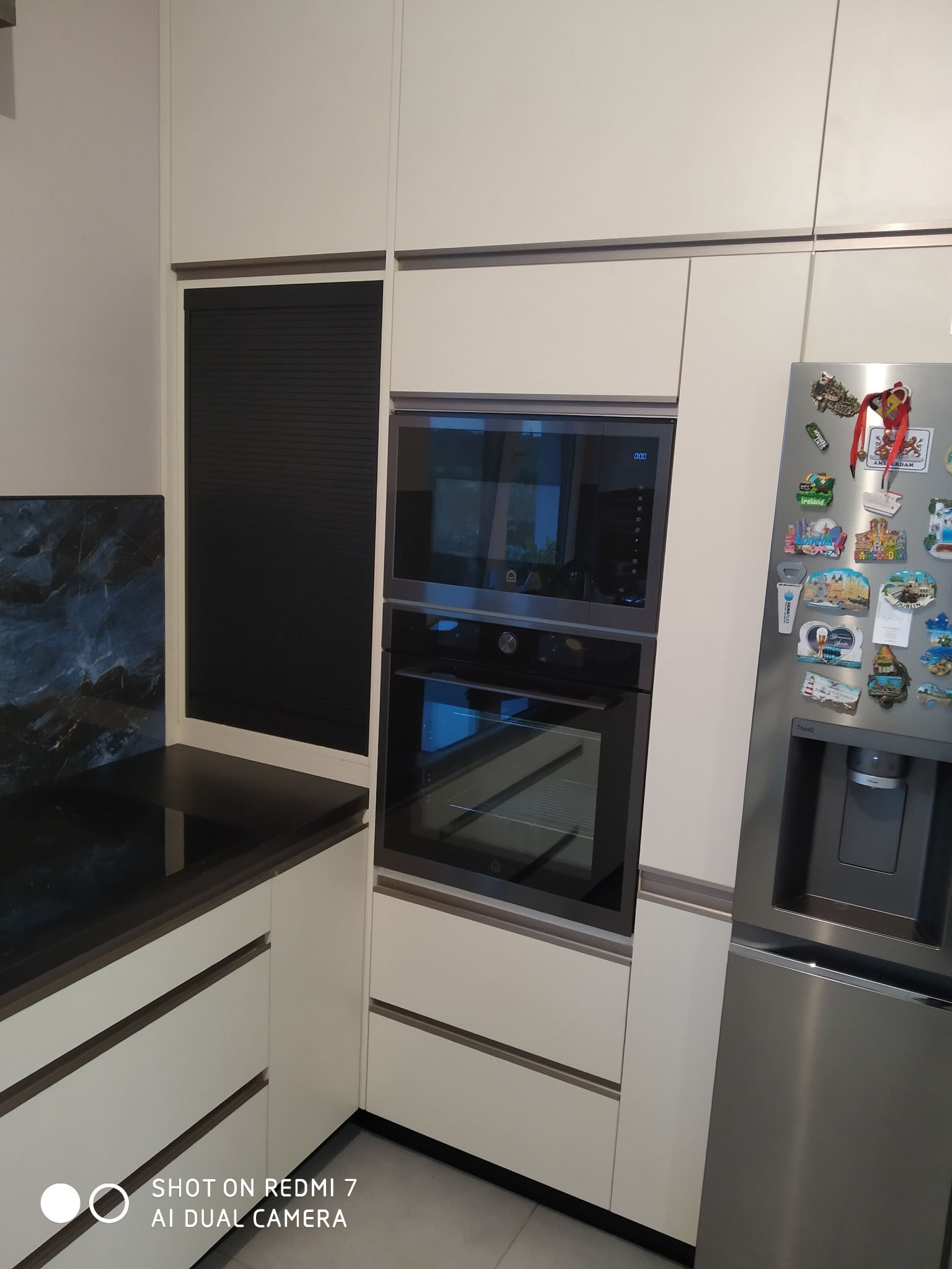 Nowoczesna zabudowa kuchenna z białymi frontami, piekarnikiem i mikrofalówką w zabudowie, czarnym blatem i panelem nad blatem imitującym marmur, oraz lodówką z magnesami.