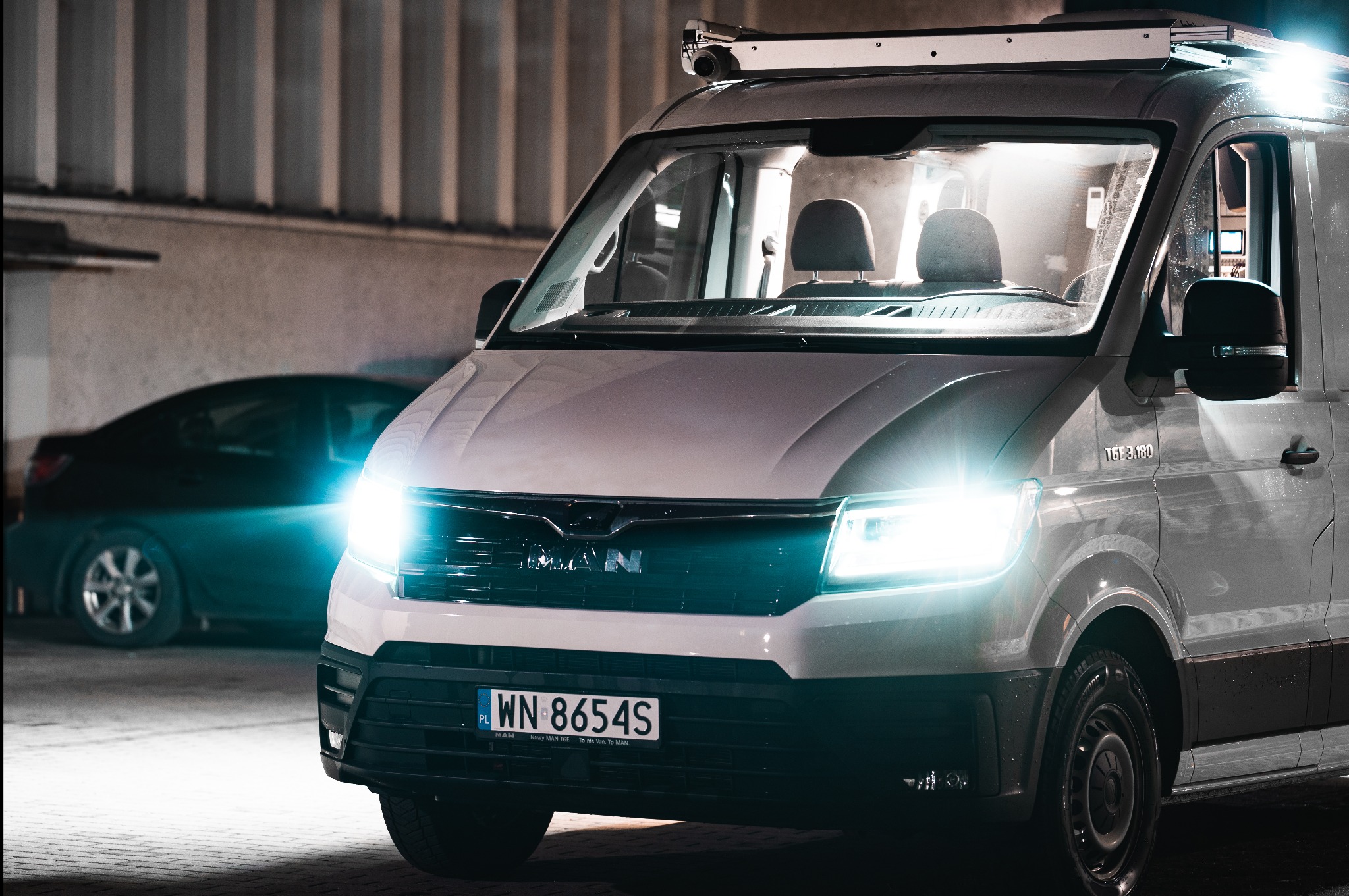 Biały van MAN TGE 3.180 z oświetleniem na dachu, zaparkowany w garażu, z widoczną tablicą rejestracyjną WN 8654S i czarnym samochodem w tle.
