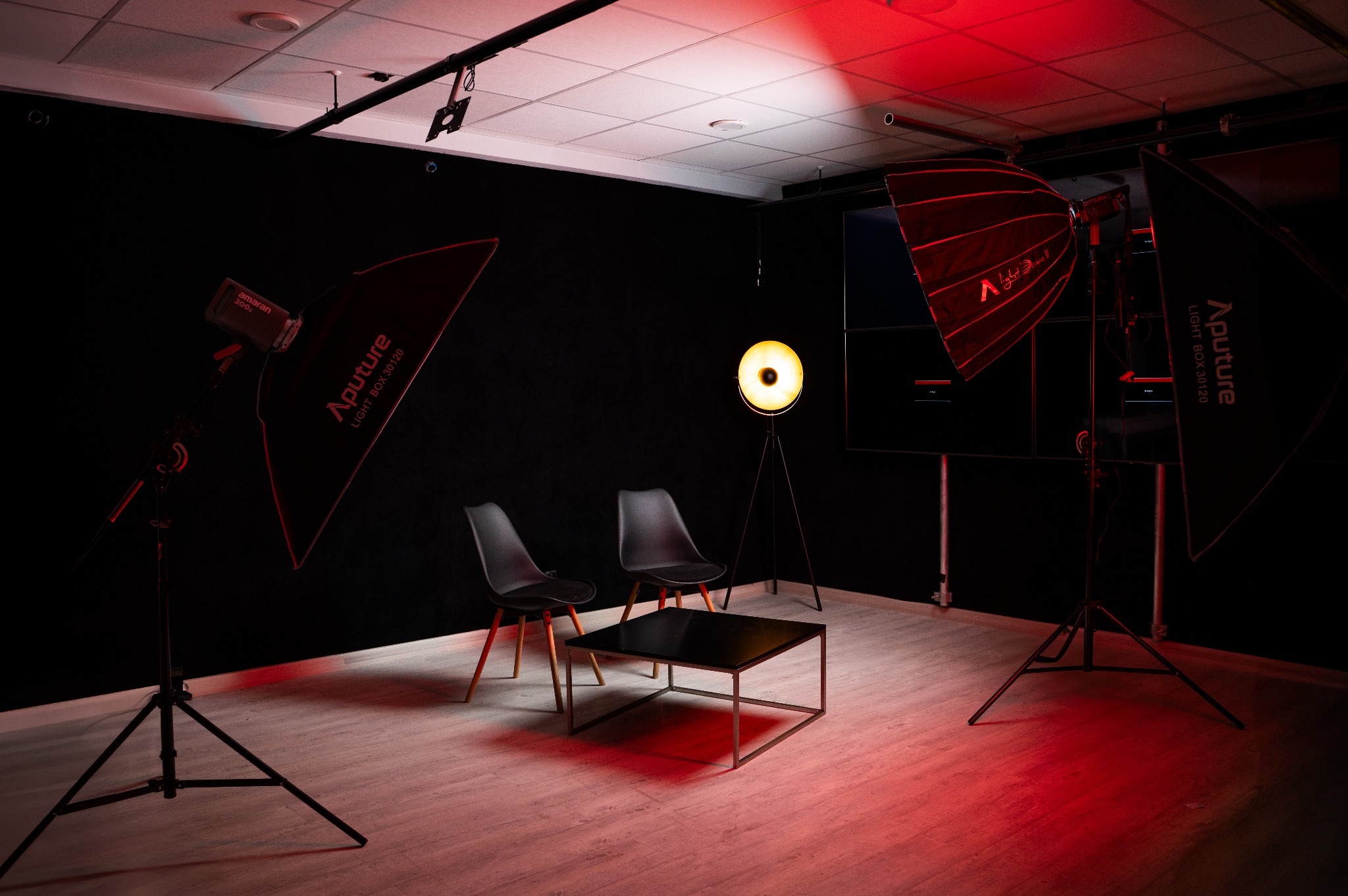 Profesjonalne studio filmowe z czarnym tłem, dwoma krzesłami, stolikiem kawowym i oświetleniem studyjnym, w tym lampami z softboxami Apurture i okrągłą lampą, oświetlone czerwonym światłem.