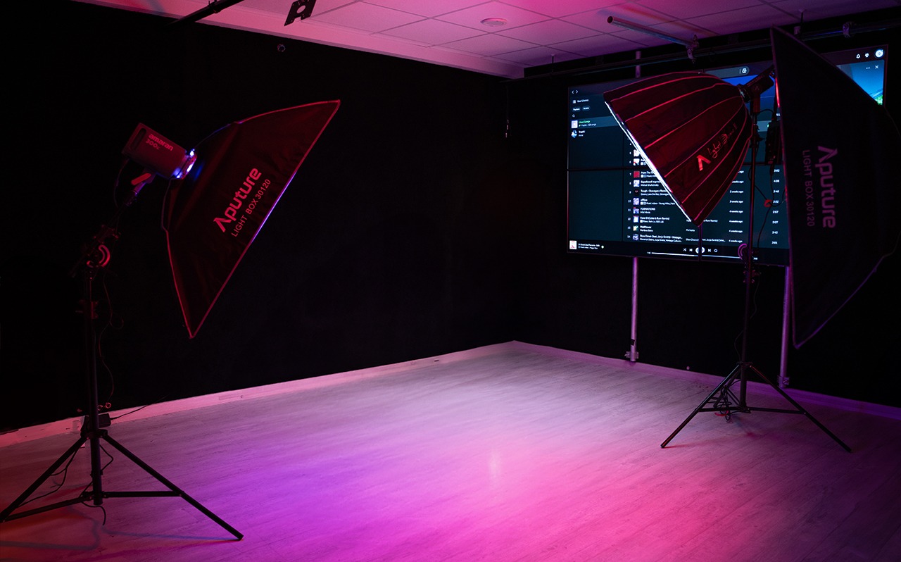 Pusta przestrzeń studia oświetlona lampami Aputure z softboxami, na ścianie widoczny duży ekran z listą odtwarzania muzyki.