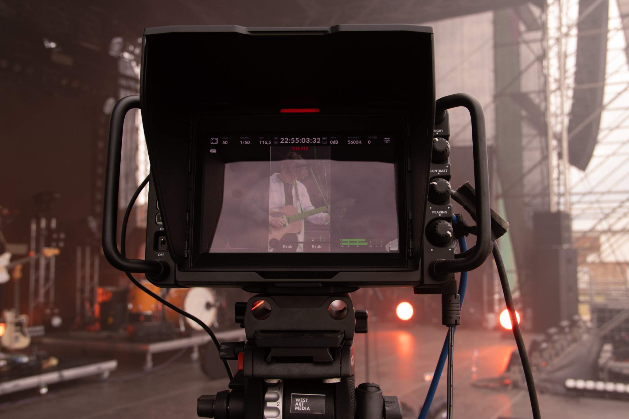 Kamera filmowa na statywie, z widokiem na ekranie na gitarzystę podczas występu na scenie, sprzęt oświetleniowy w tle.