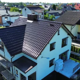 Dom z ciemnobrązowym, błyszczącym dachem, widoczny z góry. Na pierwszym planie rusztowanie. W tle inne domy i zieleń. Słoneczny dzień.