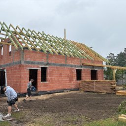 Bączkowski team „Jakub Bączkowski” - Budowa domu z czerwonej cegły. Widoczna więźba dachowa w trakcie montażu, deski, drabina i pracownicy. Pochmurne niebo w tle, ziemia po deszczu.