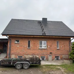 Dom z czerwonej cegły z nowym, ciemnym dachem z dachówki ceramicznej. Przyczepa z materiałami budowlanymi na posesji. Widoczne elementy wykończenia dachu.