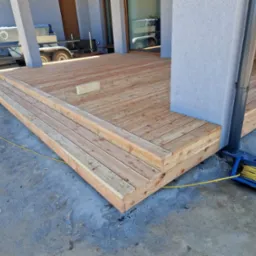 Drewniany taras z trzema stopniami, świeżo po montażu, widoczny zwój żółtego kabla elektrycznego i przyczepka w tle.