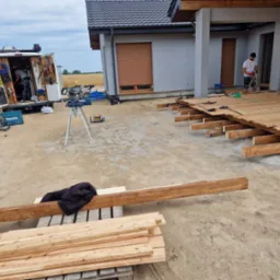Montaż drewnianego tarasu przy domu jednorodzinnym, widoczne deski, legary, narzędzia i pracownik w trakcie pracy.