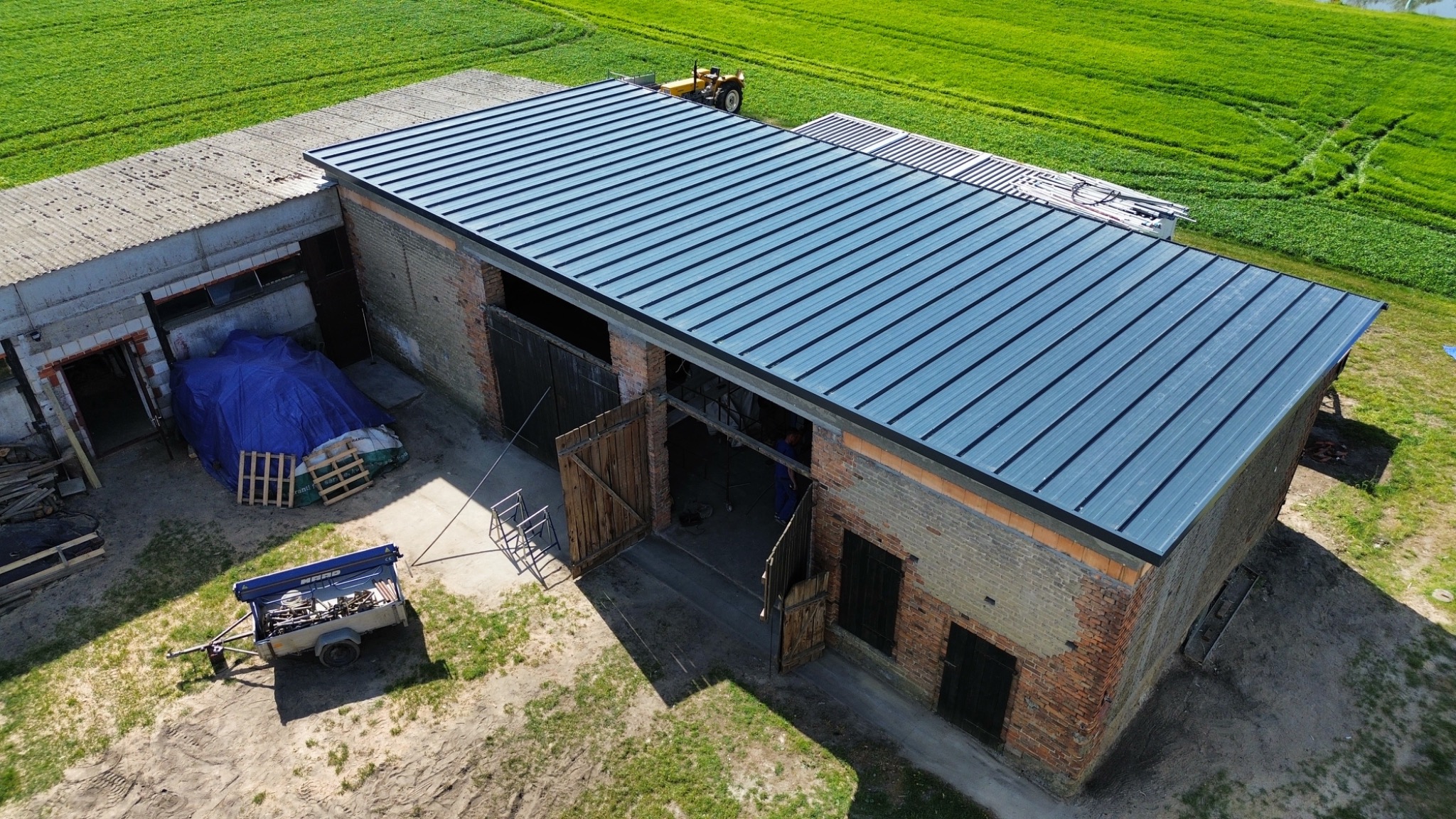 Widok z lotu ptaka na budynek gospodarczy z nowym, metalowym pokryciem dachowym w odcieniach szarości, z widocznym traktorem na tle zielonego pola.