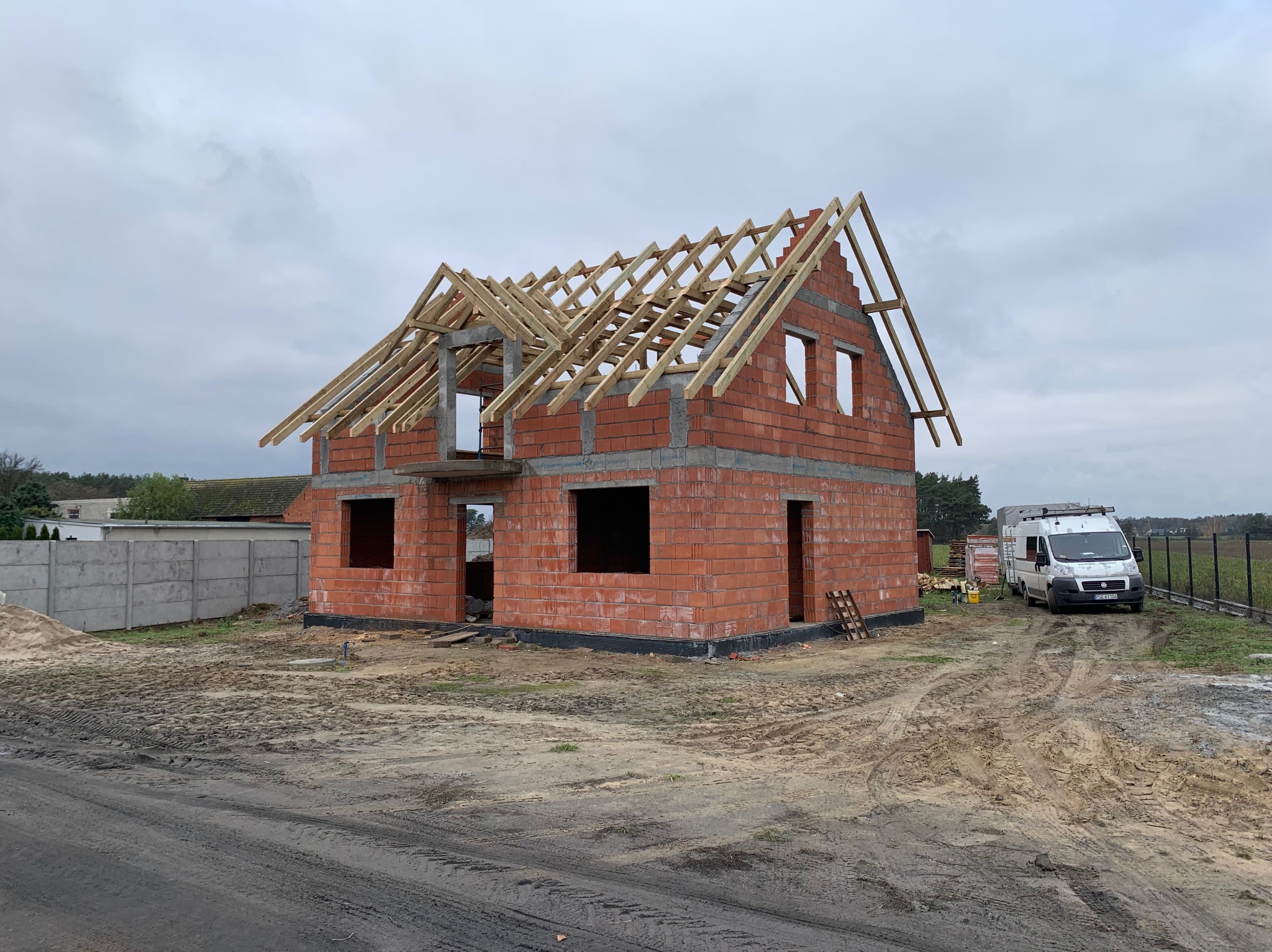 Surowy stan domu w trakcie budowy: czerwona cegła, drewniana więźba dachowa, widoczny samochód dostawczy na posesji, teren wokół domu w stanie przygotowania.