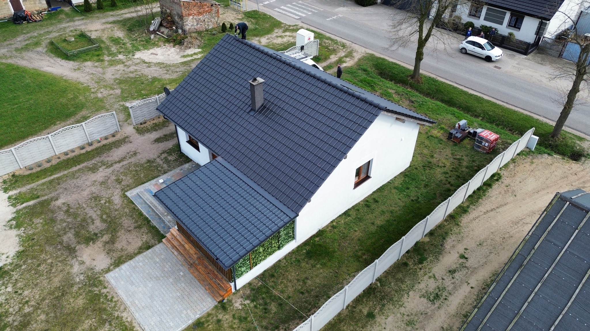 Widok z góry na dom jednorodzinny z nowym dachem z ciemnej dachówki ceramicznej, kominem i drewnianym tarasem, otoczony ogrodzeniem betonowym i zielenią.