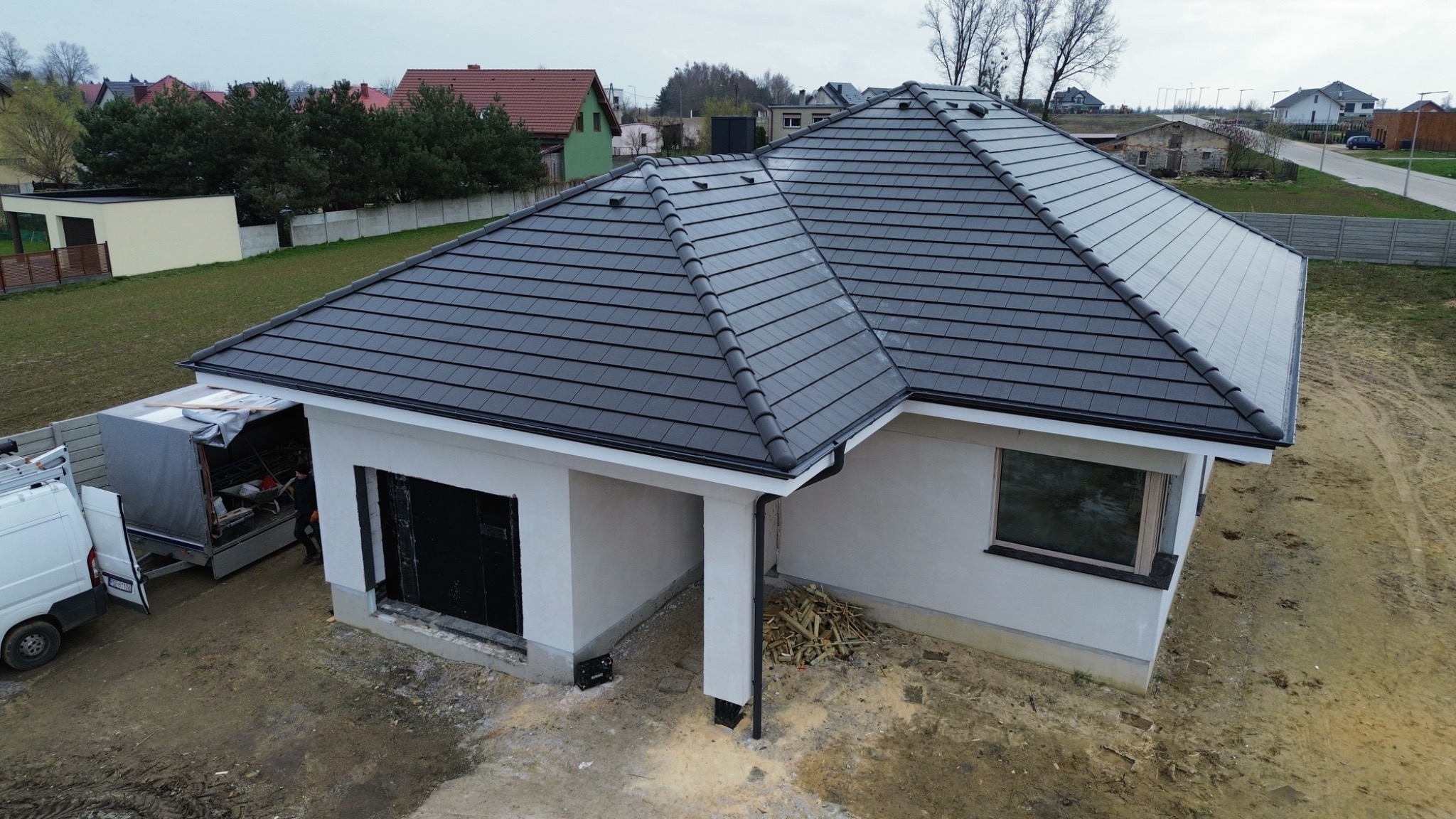 Nowoczesny dom jednorodzinny z grafitową dachówką modułową, widok z góry, na podwórku leżą resztki drewna, w tle zabudowa jednorodzinna.