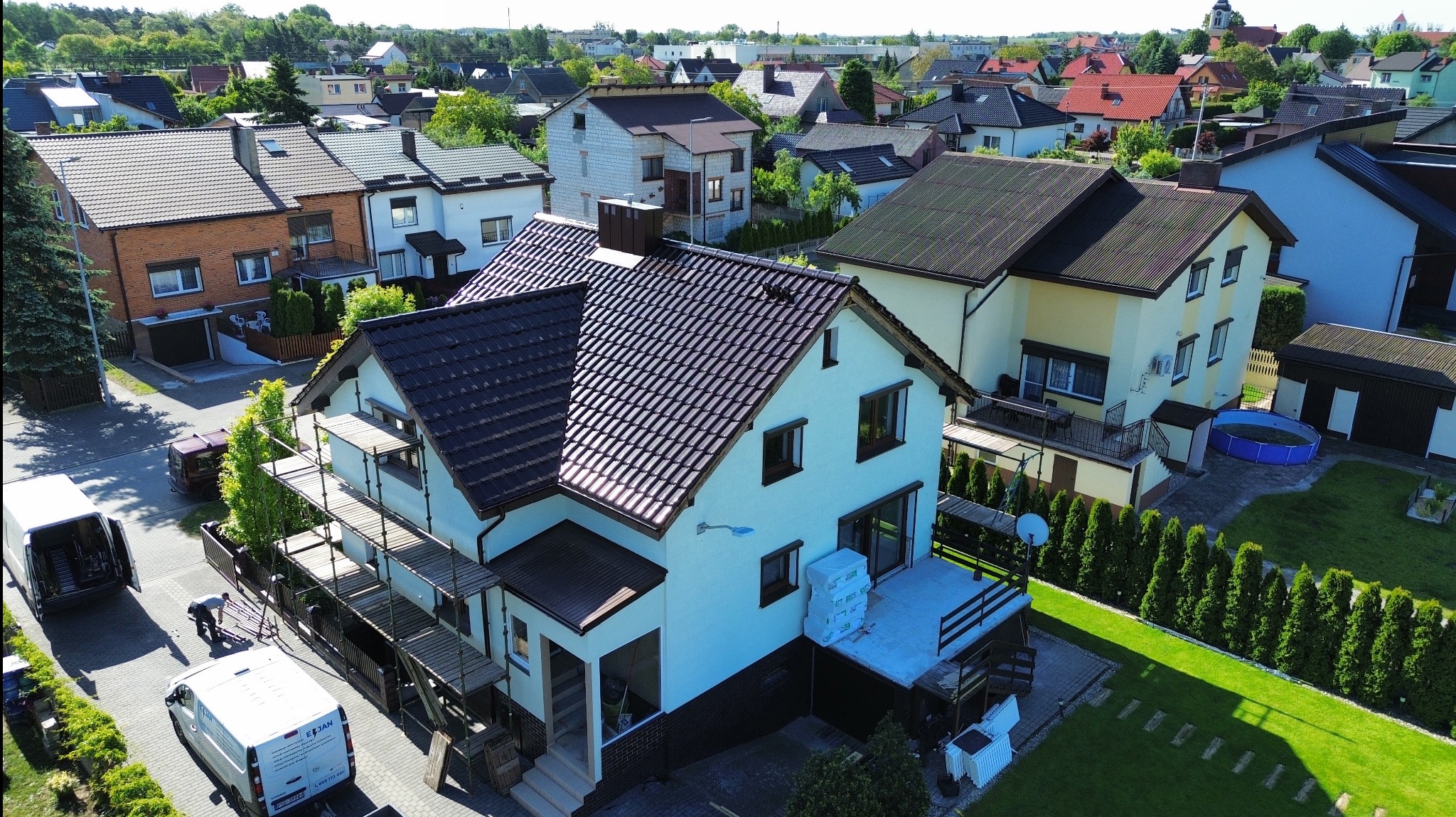 Dom z brązowym dachem w trakcie prac dekarskich, widoczne rusztowania i materiały budowlane. Ujęcie z góry, w tle inne domy i zieleń.