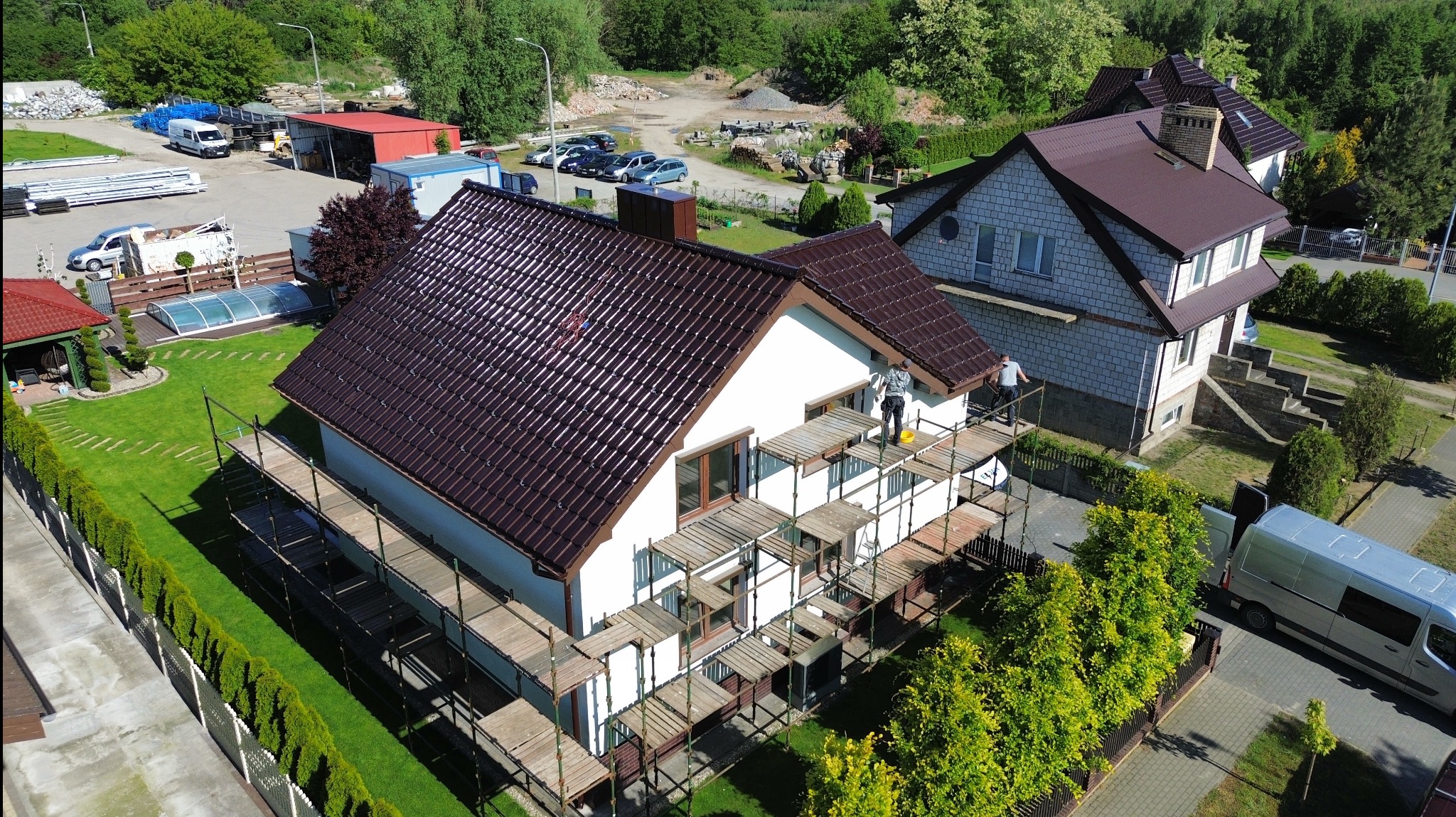 Montaż brązowej dachówki na białym domu z rusztowaniem. Widok z góry ukazuje dom, otoczenie i pracowników przy pracy.