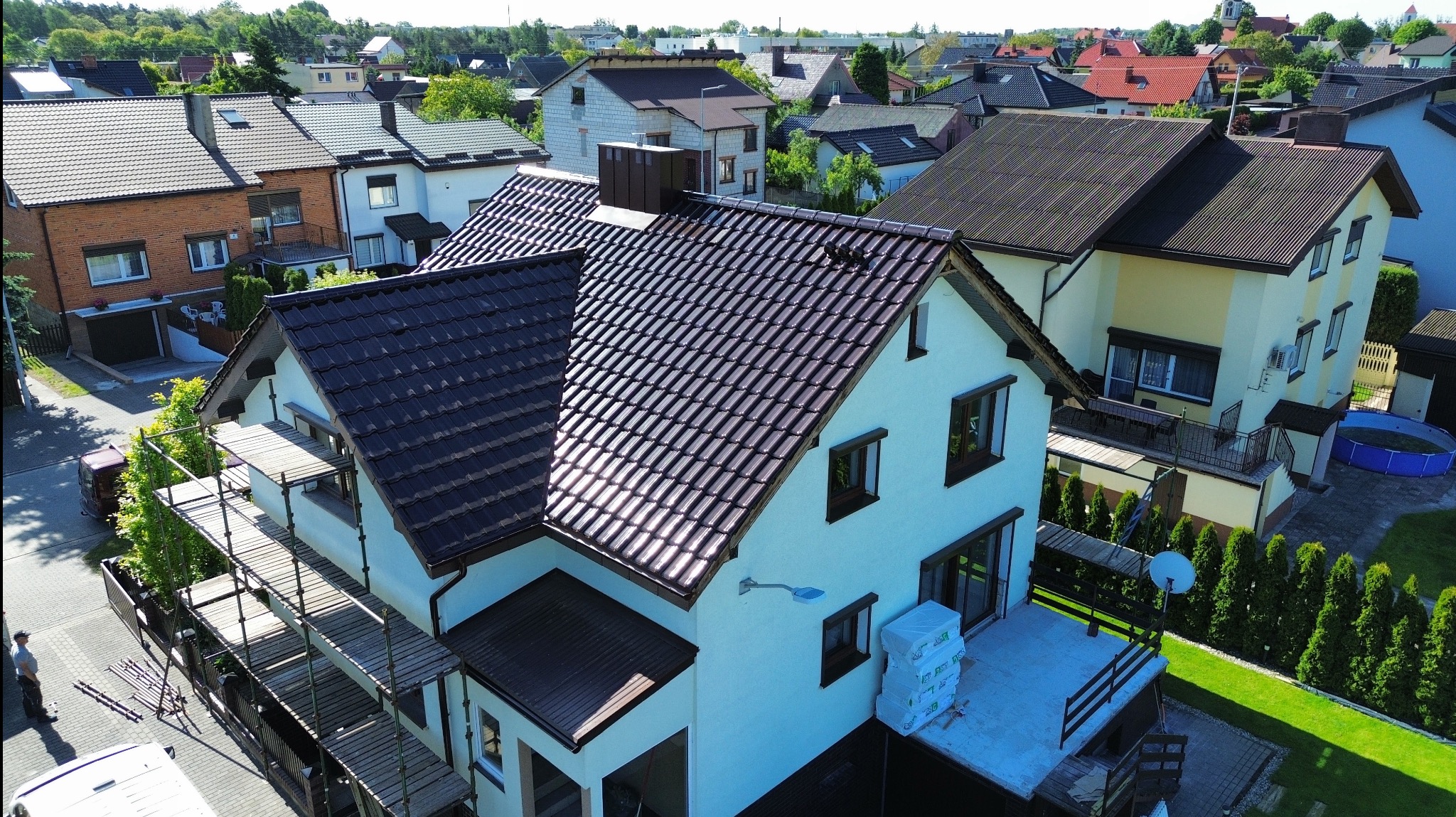 Dom z ciemnobrązowym, błyszczącym dachem, widoczny z góry. Na pierwszym planie rusztowanie. W tle inne domy i zieleń. Słoneczny dzień.