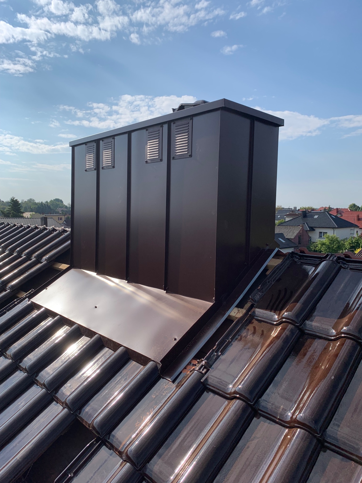 Nowy, brązowy komin wentylacyjny na błyszczącej dachówce ceramicznej w słoneczny dzień. Widoczne detale montażu i okolica z domami.