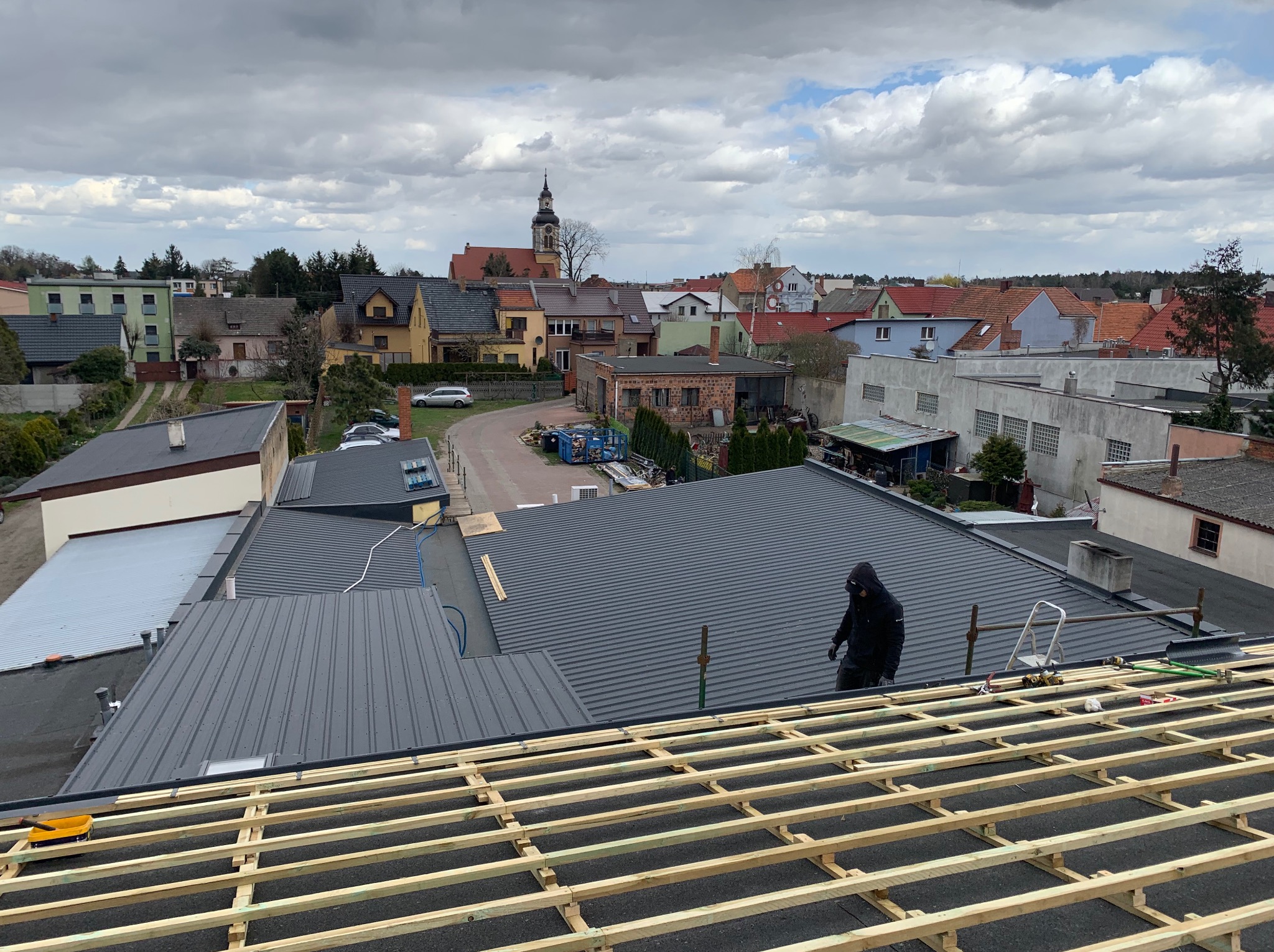 Montaż blachy trapezowej na dachu budynku. Widoczna więźba dachowa, narzędzia i częściowo ukończone krycie. W tle zabudowa miejska i pochmurne niebo.