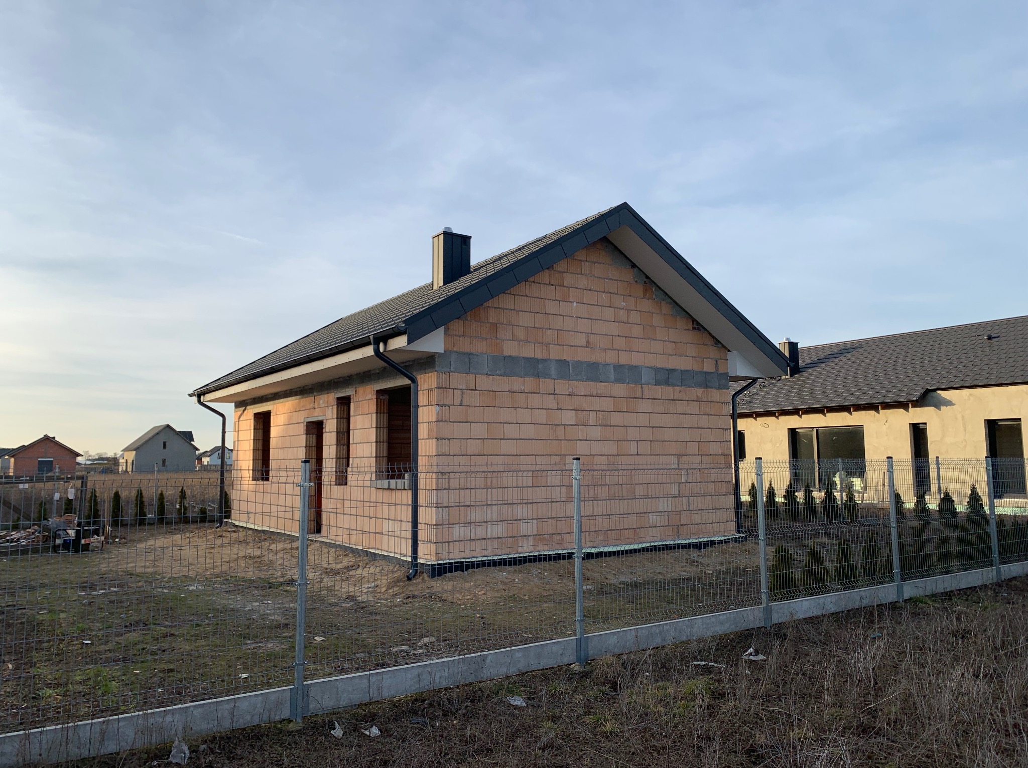 Surowy stan domu z dachem w Jarocinie, widok z zewnątrz, ogrodzenie siatkowe. Budynek w trakcie budowy z cegły, czarny dach i komin, okna bez szyb.