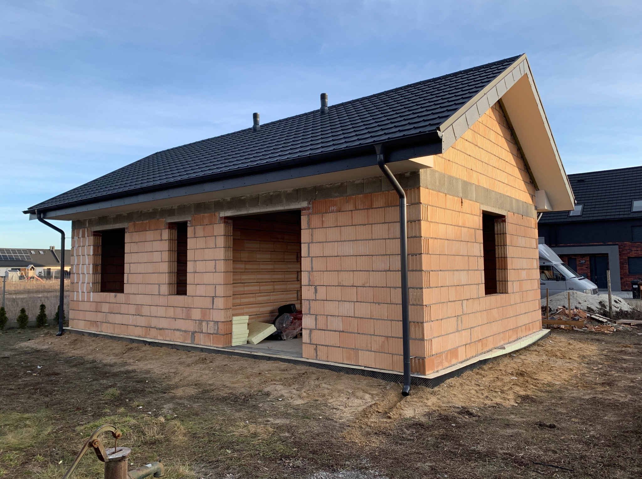 Surowy budynek jednorodzinny z ciemnym dachem z dachówki, widoczne otwory okienne i drzwiowe, oraz orynnowanie. Budowa w trakcie realizacji.