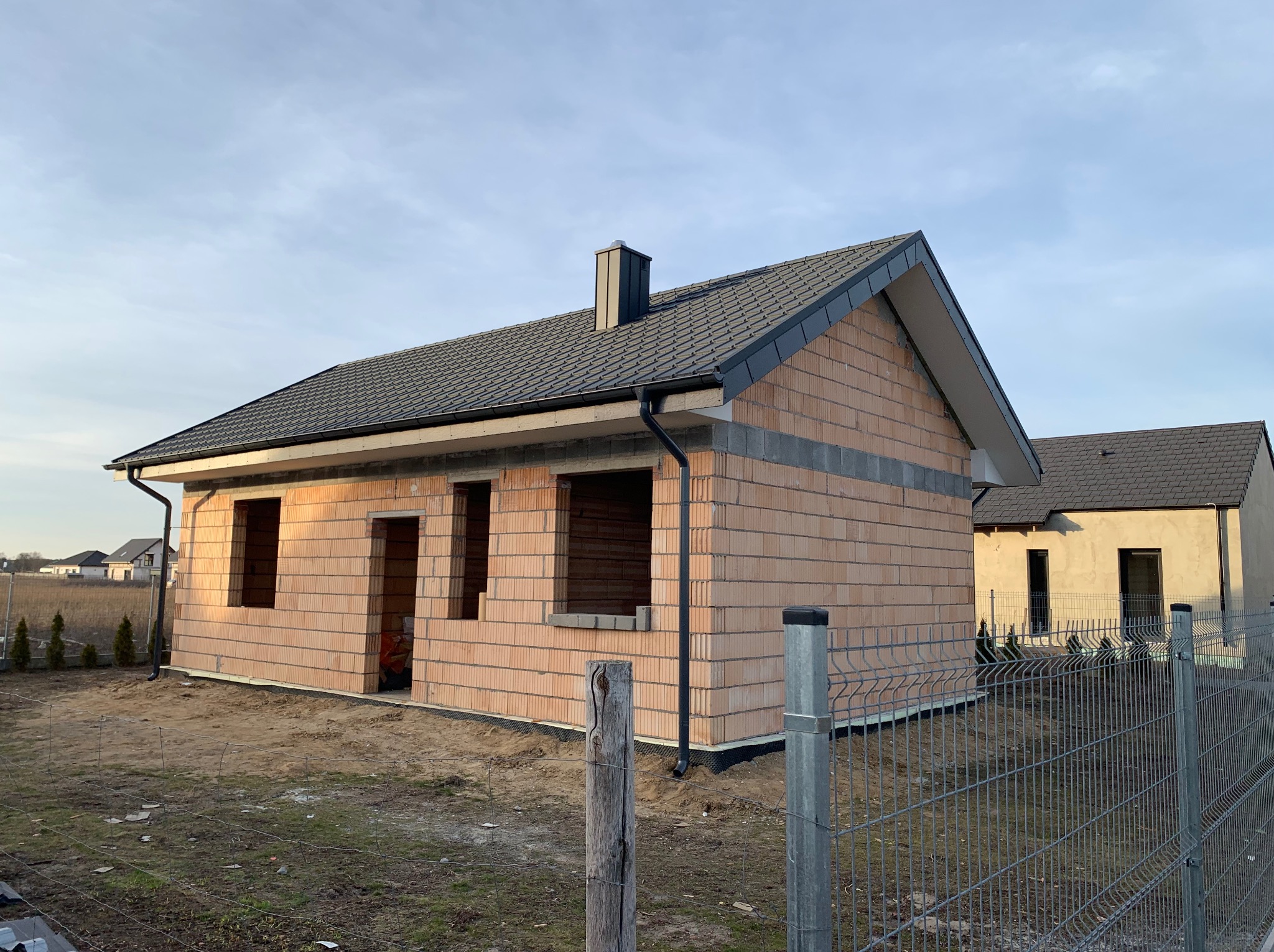 Dom w trakcie budowy z widoczną konstrukcją dachu i ścianami z cegły ceramicznej. W tle drugi budynek i błękitne niebo.