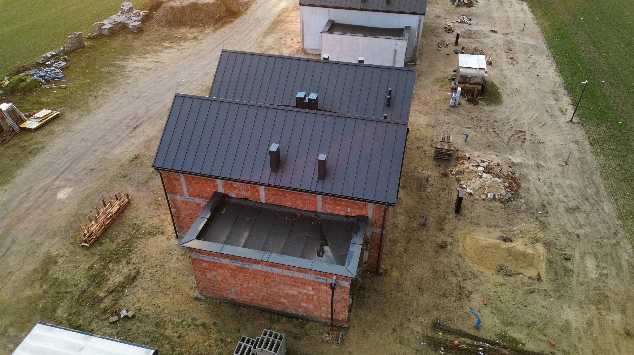 Dom w trakcie budowy z ciemnym dachem blaszanym, widok z góry, surowe otoczenie budowy, ceglana konstrukcja wznoszona, teren przygotowany pod budowę, widoczne materiały budowlane.