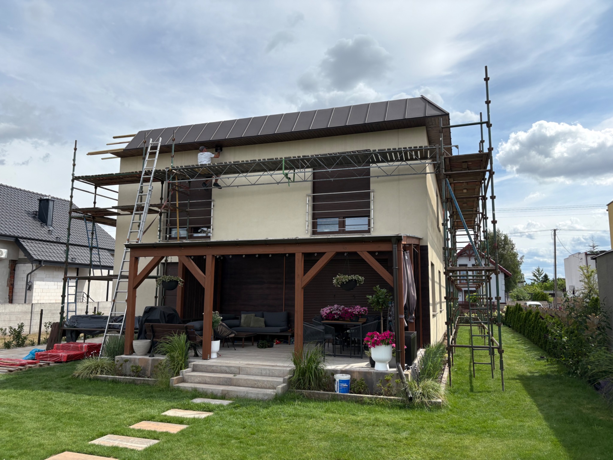 Dom jednorodzinny w trakcie krycia dachu brązową blachodachówką, widoczne rusztowania i dekarz podczas pracy. W tle zielony ogród.