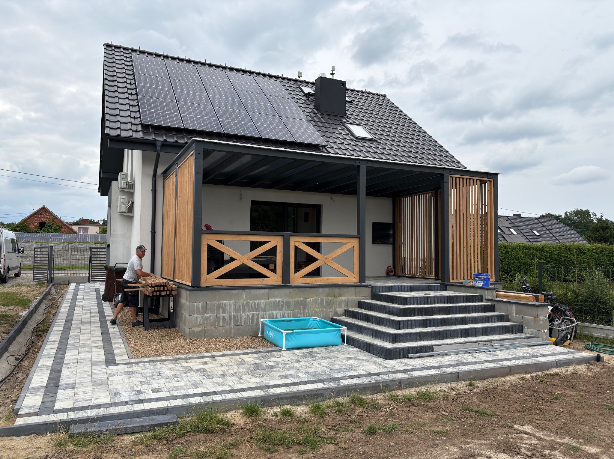 Nowoczesny dom z panelami słonecznymi i zadaszonym tarasem. Ułożona kostka brukowa na podjeździe, pracownik w trakcie układania drewnianych elementów tarasu.