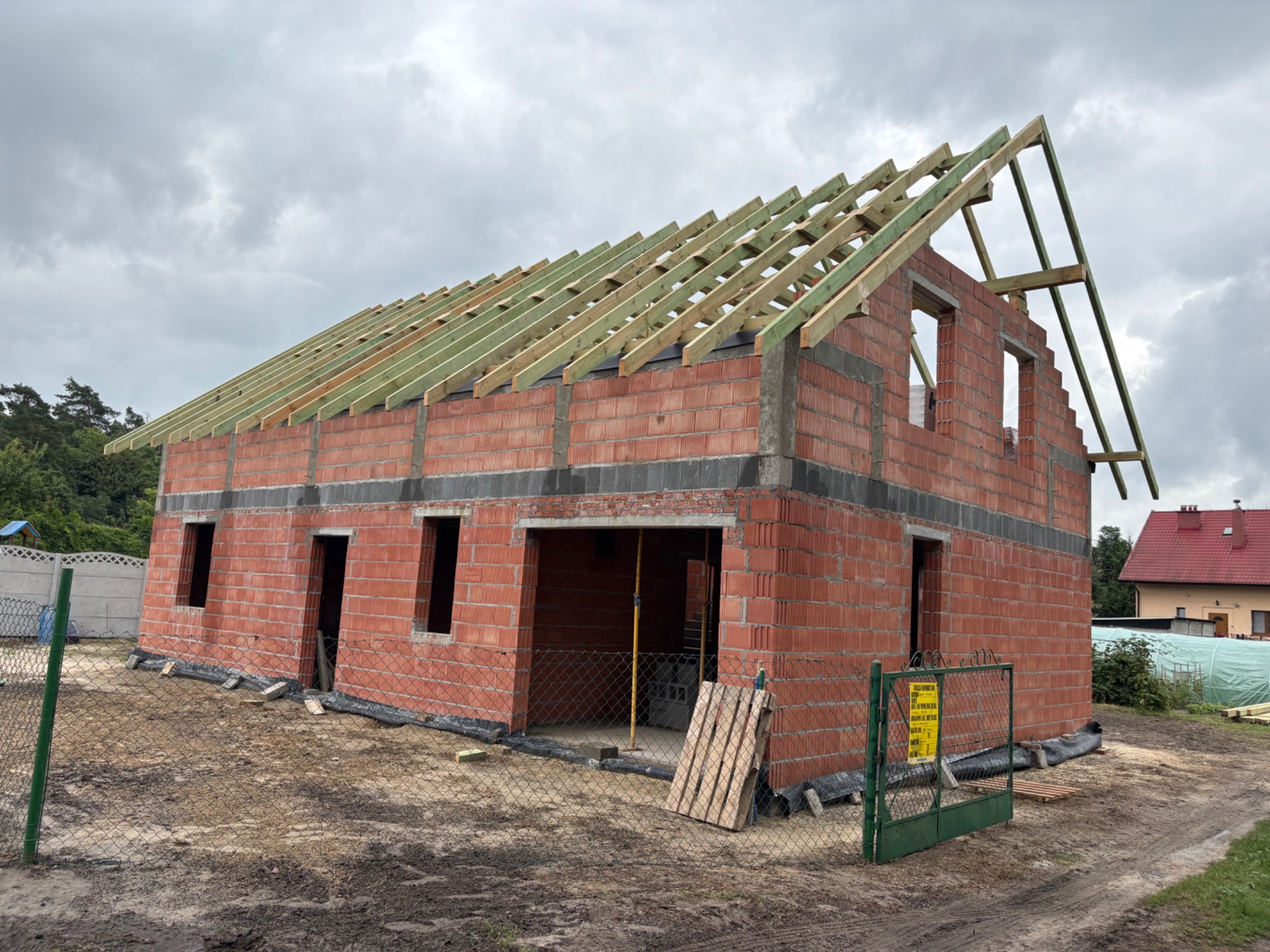 Surowy budynek z czerwonej cegły w trakcie budowy więźby dachowej. Widoczne drewniane elementy konstrukcyjne i niezadaszone okna. Na pierwszym planie ogrodzenie z siatki.