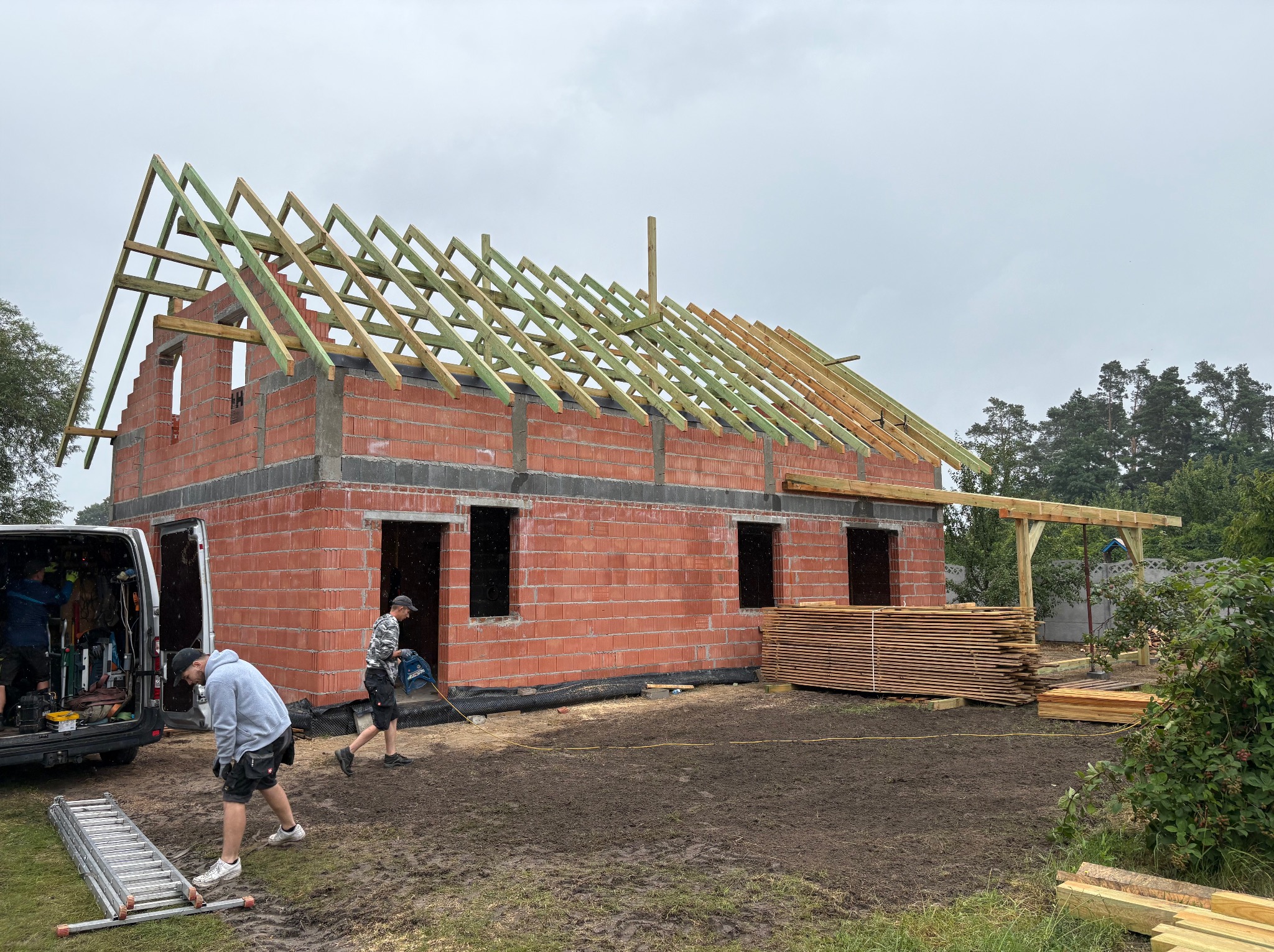 Budowa domu z czerwonej cegły. Widoczna więźba dachowa w trakcie montażu, deski, drabina i pracownicy. Pochmurne niebo w tle, ziemia po deszczu.