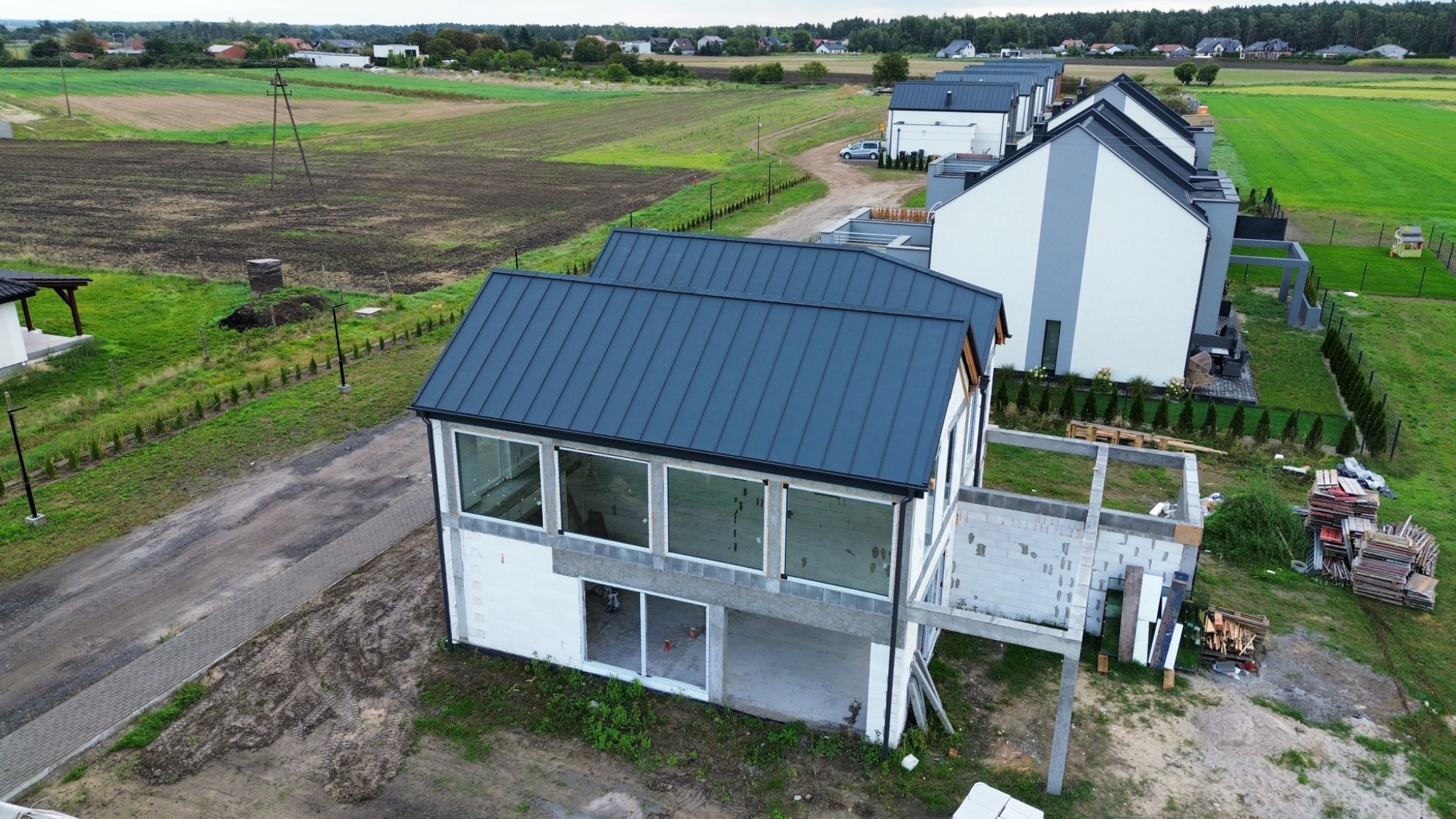 Nowoczesny dom w trakcie budowy z ciemnoszarym dachem na tle krajobrazu. Widoczne elementy konstrukcyjne i otoczenie budowy z paletami materiałów.