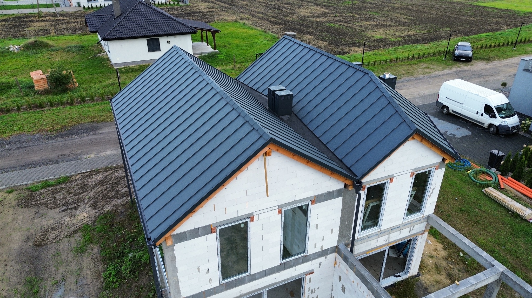 Nowoczesny, grafitowy dach z blachy na domu w stanie surowym otwartym, widok z góry. W tle drugi dom z identycznym dachem i pole uprawne.