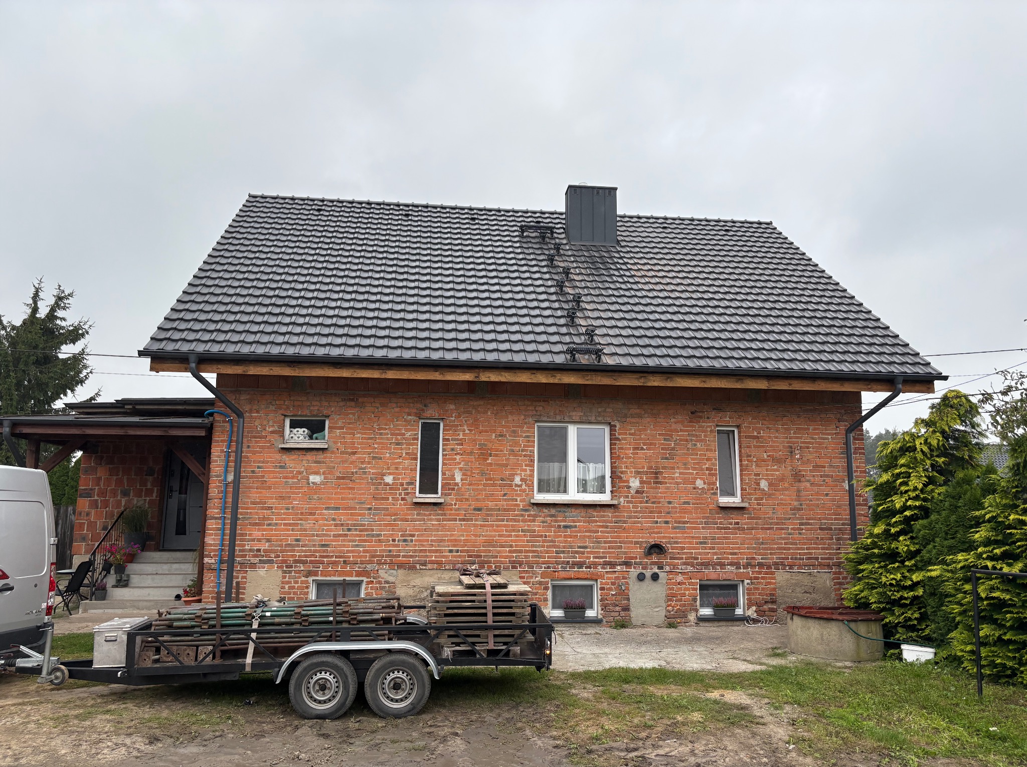 Dom z czerwonej cegły z nowym, ciemnym dachem z dachówki ceramicznej. Przyczepa z materiałami budowlanymi na posesji. Widoczne elementy wykończenia dachu.