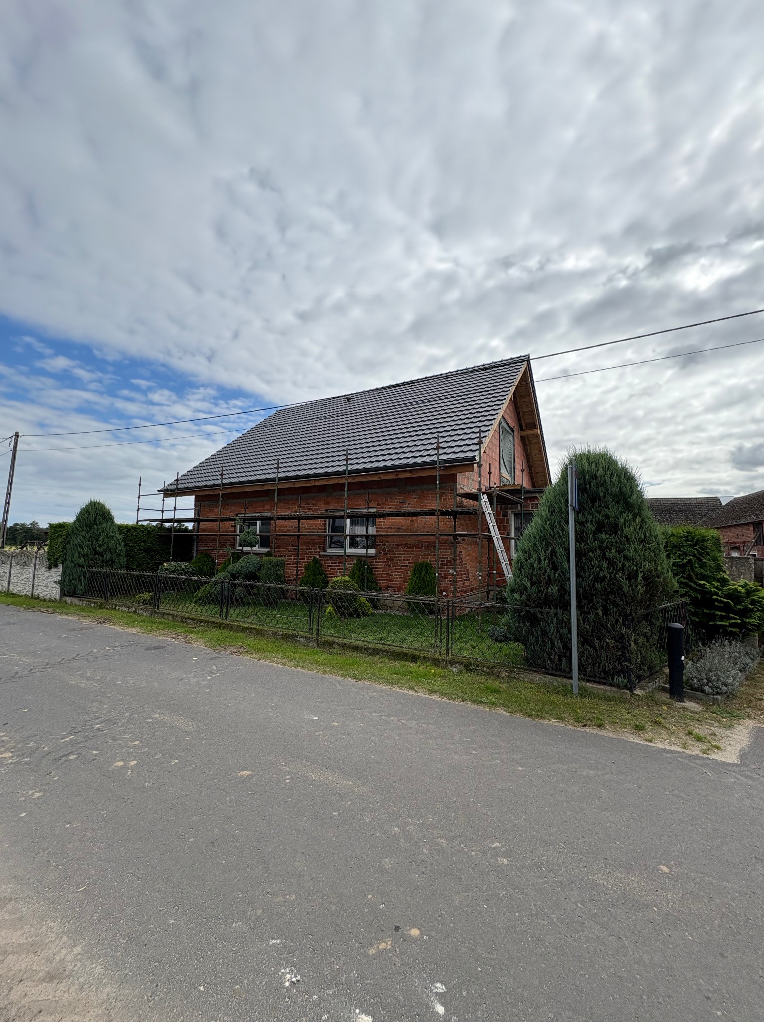 Dom z czerwonej cegły w trakcie krycia dachu szarą dachówką, otoczony rusztowaniami i zielenią, widok z ulicy w pochmurny dzień.