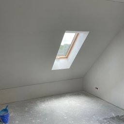 Usługi Budowlane Marek Łukojć - Poddasze po nałożeniu gładzi, widoczne ślady po pracy na podłodze, niebieskie wiadro z narzędziami w rogu, okno dachowe wpuszczające światło.