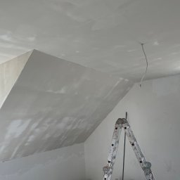 Usługi Budowlane Marek Łukojć - Poddasze po nałożeniu gładzi szpachlowej, widoczny fragment okna, kabel zwisający z sufitu oraz aluminiowa drabina oparta o ścianę.